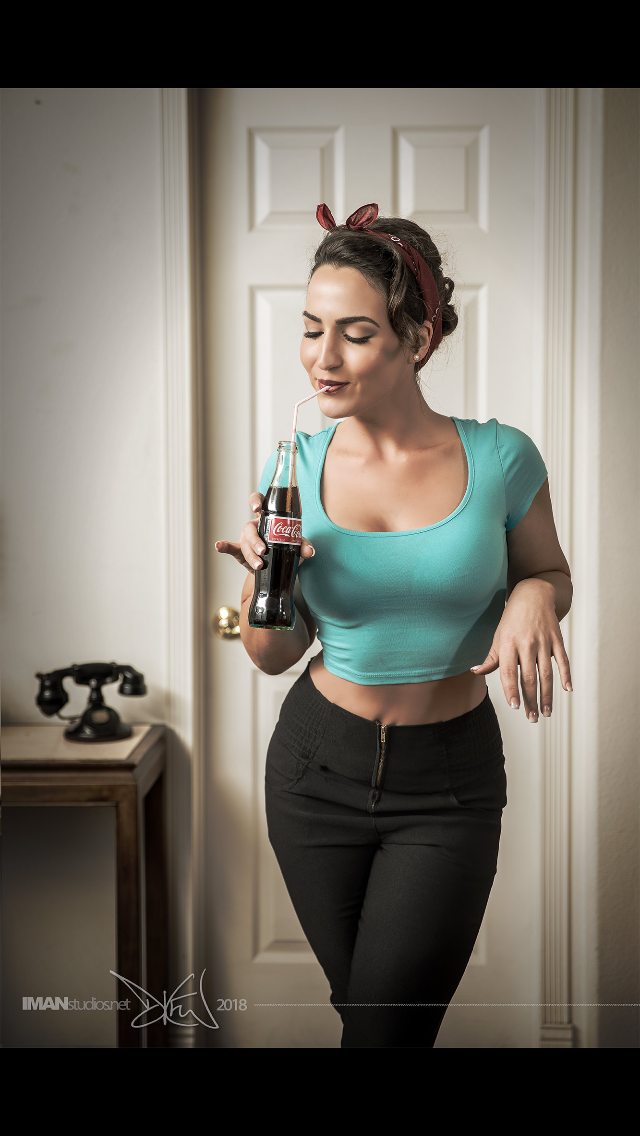 A young woman with dark hair styled in an updo, wearing a red headband, a turquoise crop top, and black high-waisted pants, drinks from a glass bottle of Coca-Cola with a straw in a room with a beige door and a vintage black rotary phone on a wooden 