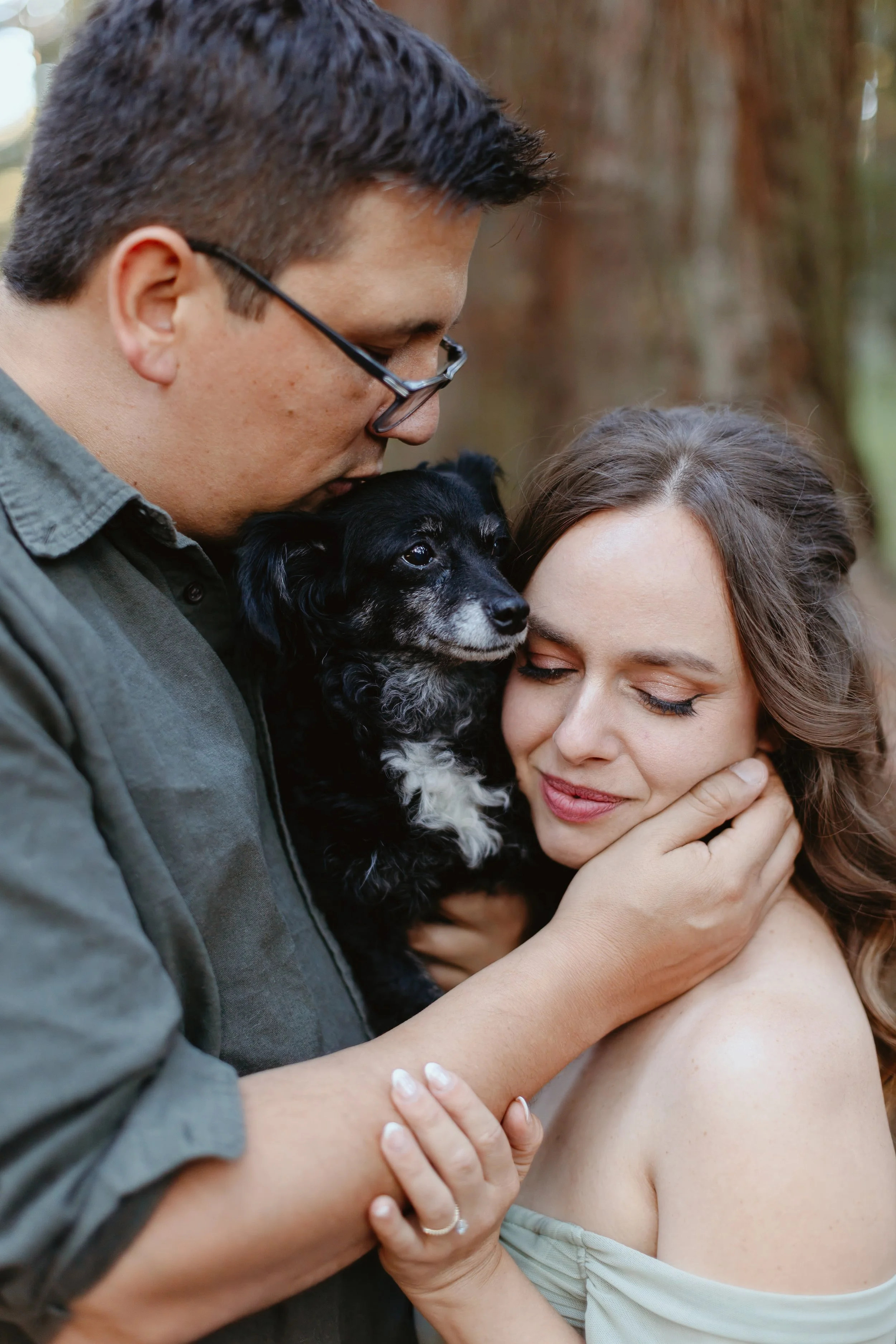 A man and woman embracing, with the man holding a black and white puppy close to his face and the woman gently holding the puppy. The man is leaning in to kiss the puppy, and the woman has her eyes closed, smiling softly.