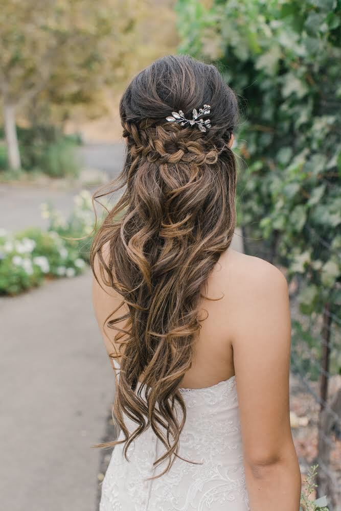 A woman with long, wavy brown hair styled with a braided crown and decorative silver hair accessory, wearing a strapless white dress, standing outdoors with blurred green foliage and flowers in the background.