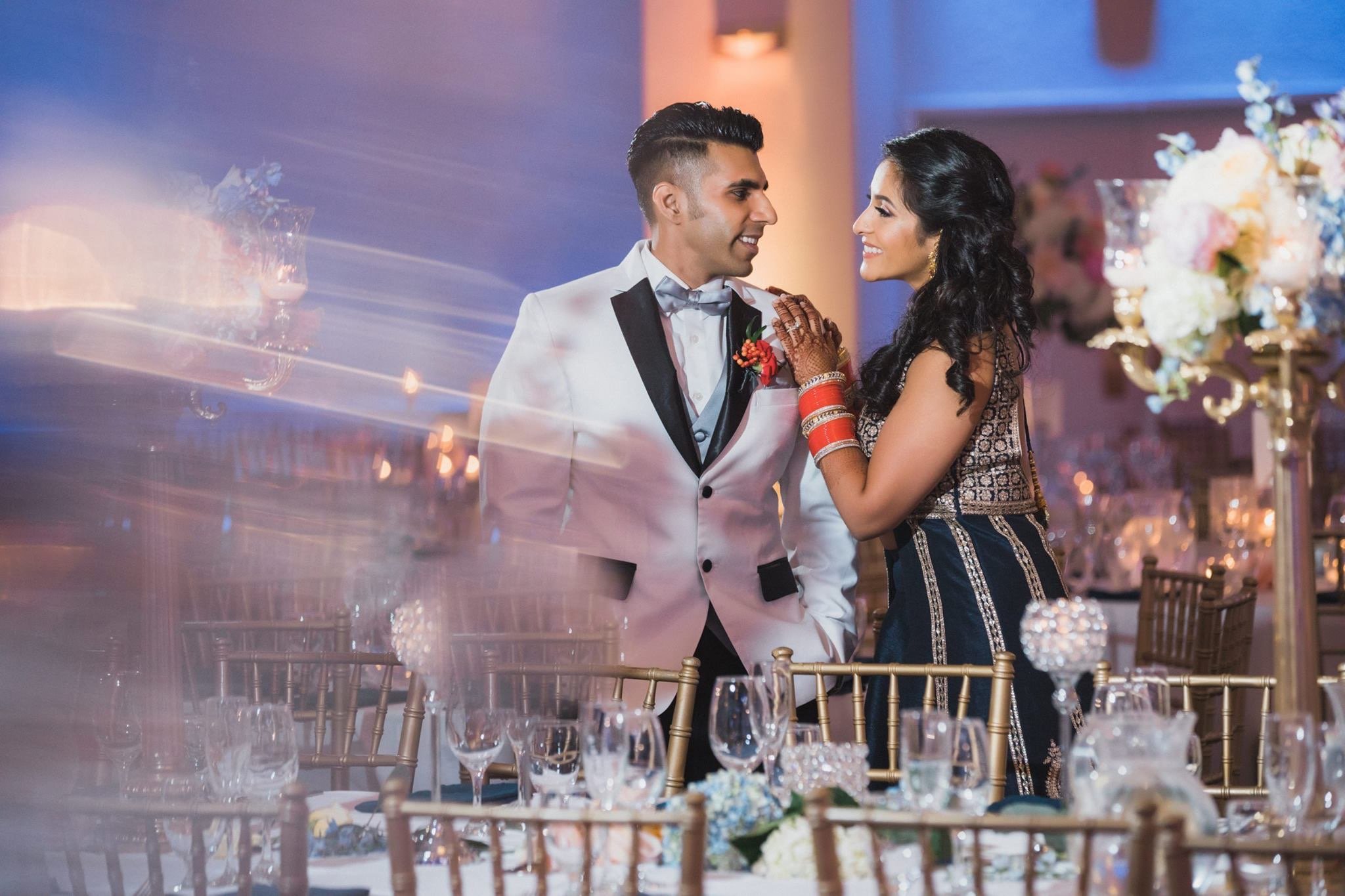 A newlywed couple sharing a moment at their wedding reception, standing close together with the bride touching the groom's shoulder. The groom is dressed in a white tuxedo with black lapels, and the bride is wearing traditional Indian attire with ban