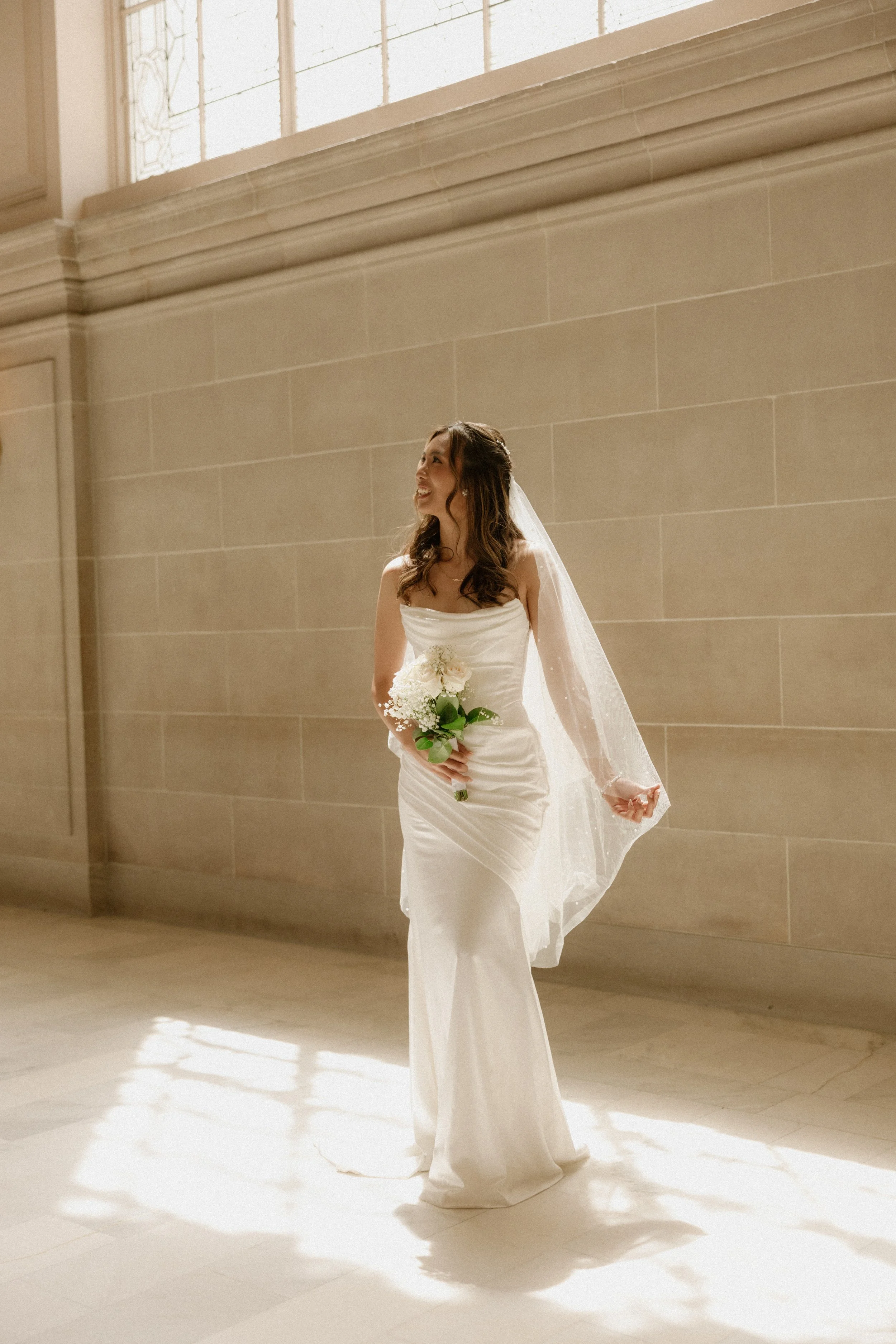 A bride in a strapless white wedding gown holding a bouquet of white flowers, standing inside a building with a large window and beige walls.