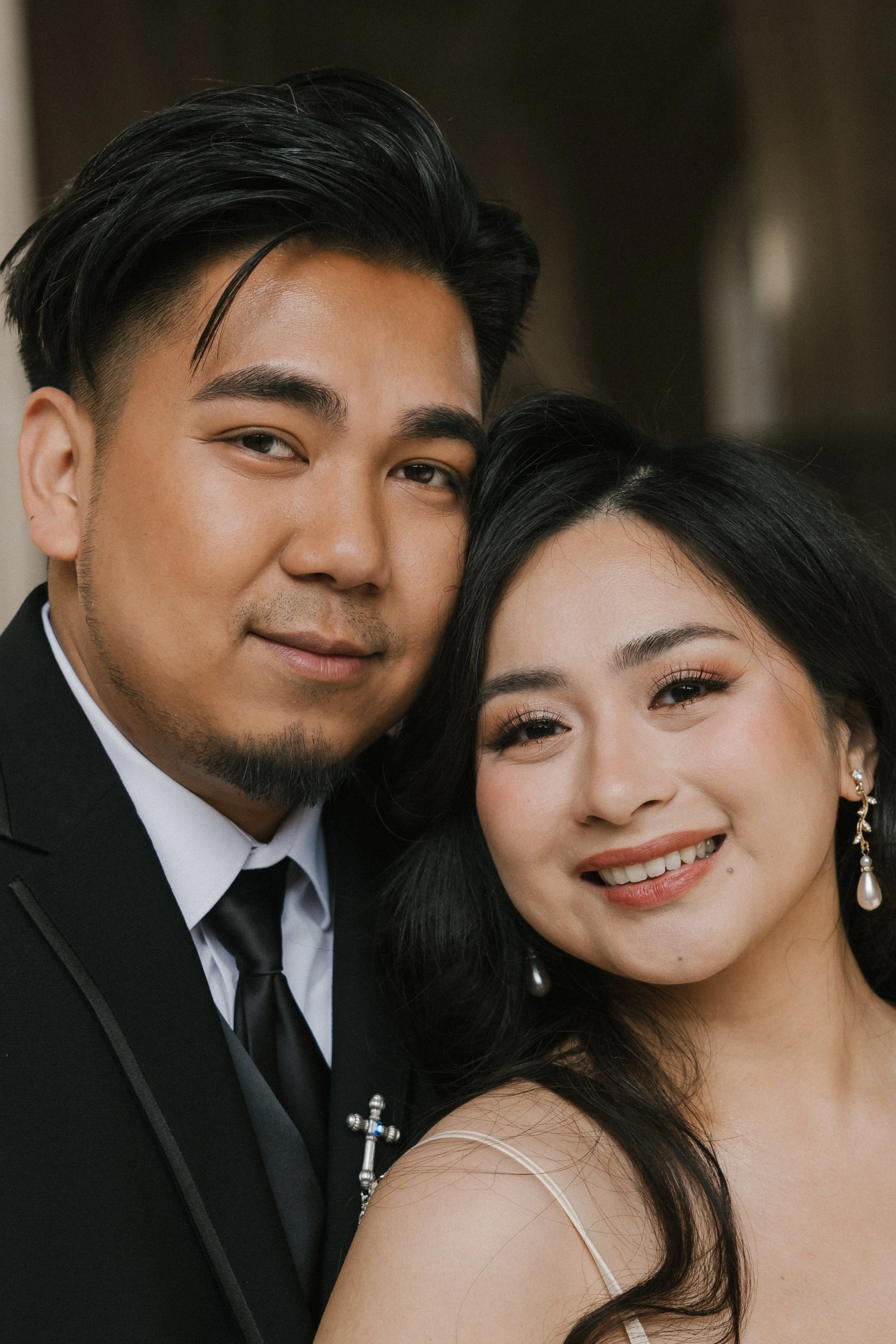 A smiling couple dressed in formal attire, the man in a black suit and tie, and the woman in earrings and a dress, standing closely together.