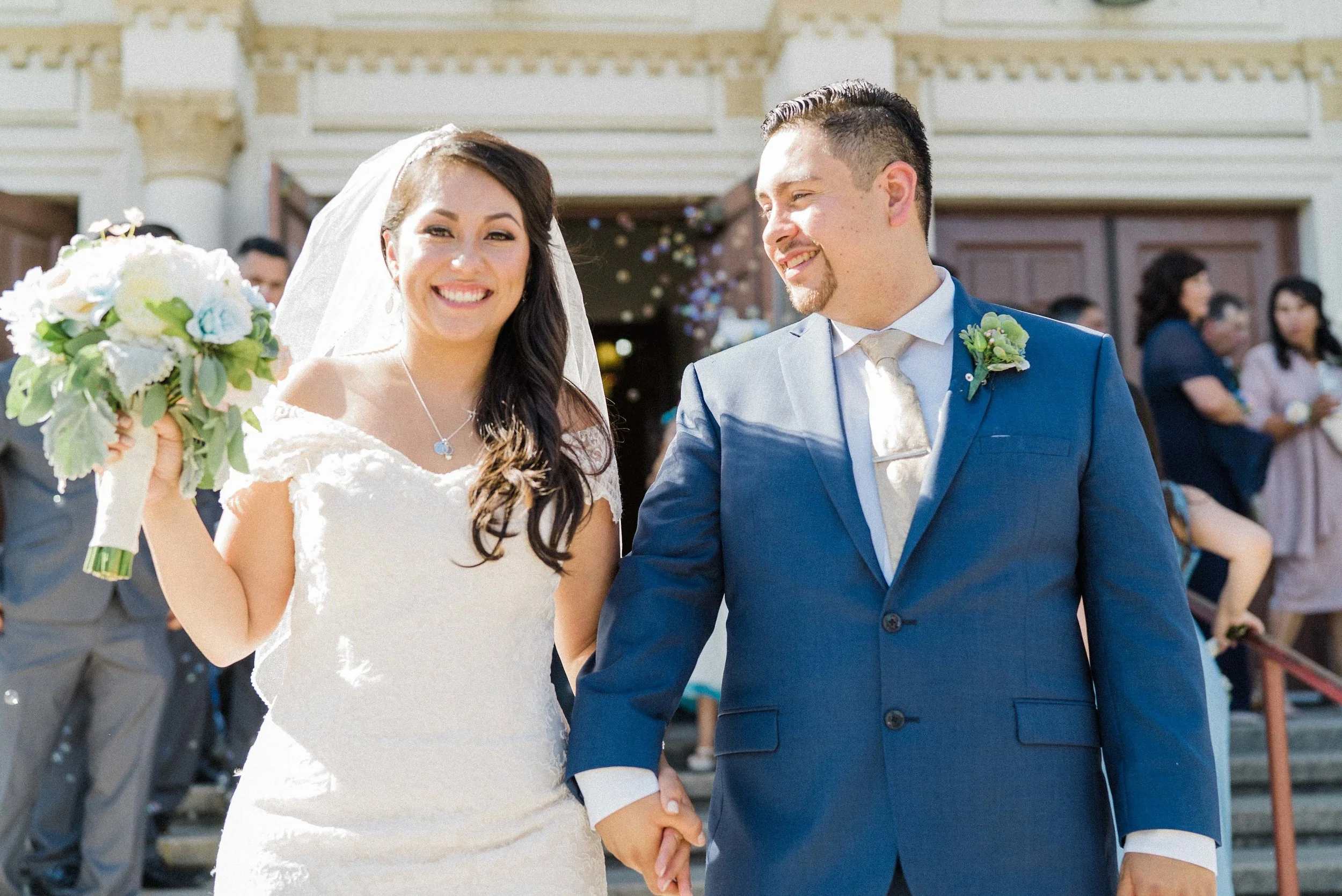Happy bride and groom holding hands and smiling outside a wedding venue, guests in the background celebrating.