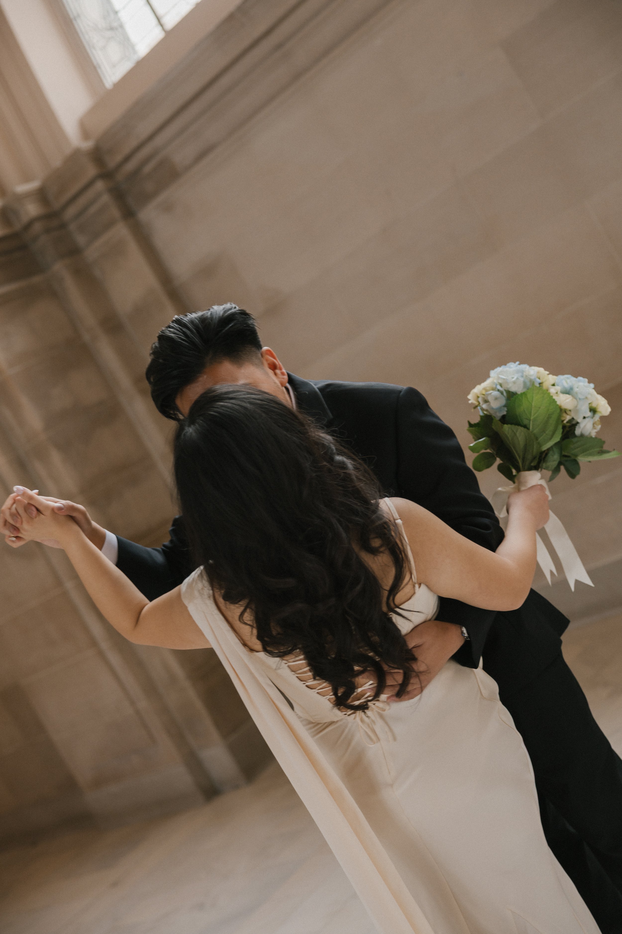 A couple dancing, with the woman holding a bouquet of white and blue flowers, in an elegant indoor setting with stone walls.