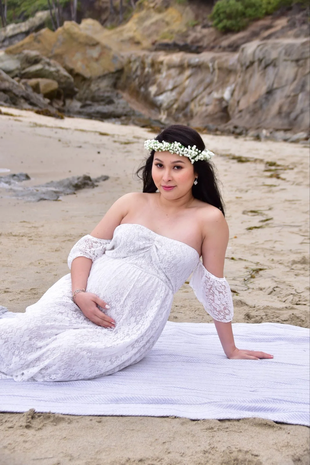 A pregnant woman in a white lace dress sitting on a towel on the sand at the beach, wearing a floral crown and earrings, with rocky cliffs and greenery in the background.