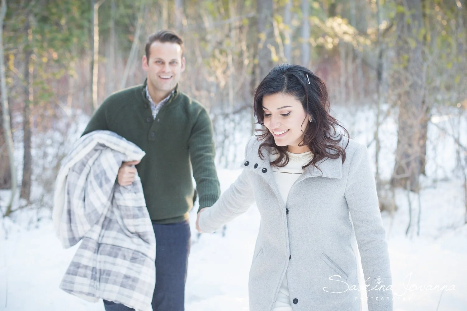 A man and woman walking in a snowy forest, holding hands, with the woman leading and smiling, and the man carrying a plaid blanket or coat.