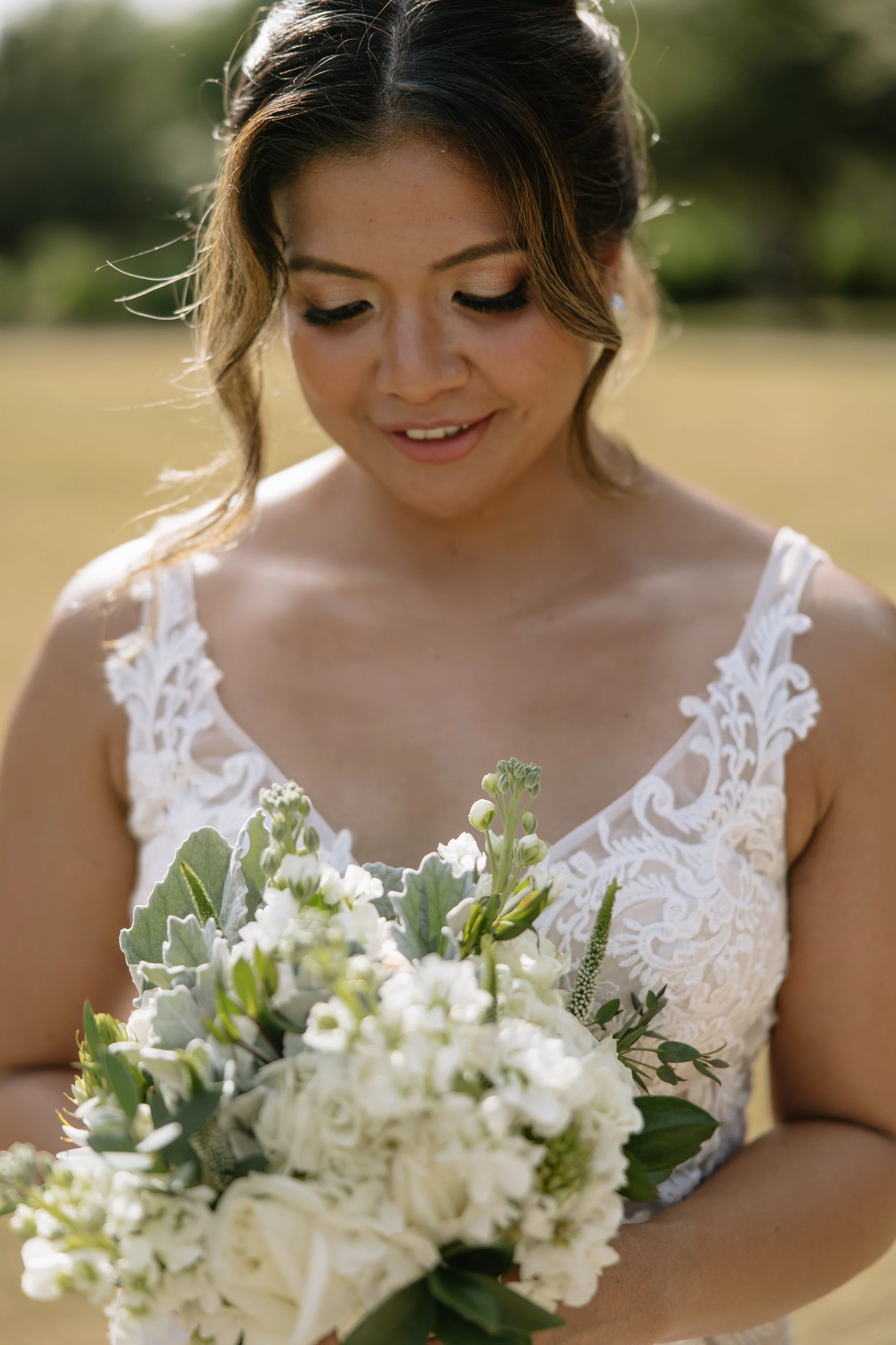 A woman in a white lace dress holding a bouquet of white flowers outdoors on a sunny day, looking down at the flowers.