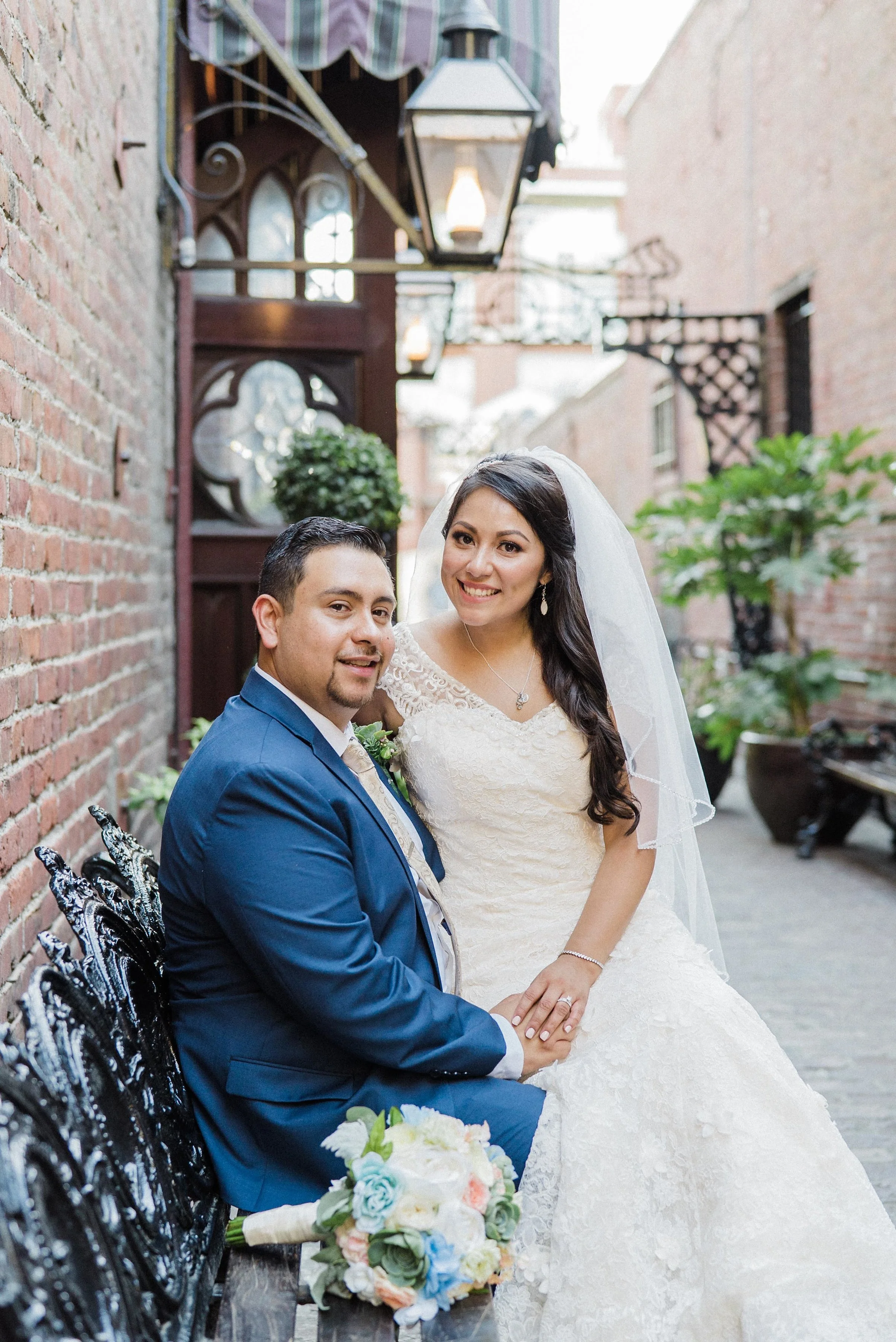A bride and groom sitting on a black ornate bench in an outdoor alleyway, smiling at the camera; the bride is wearing a white lace wedding dress with a veil, and the groom is dressed in a blue suit with a boutonniere; the bride's bouquet, consisting 