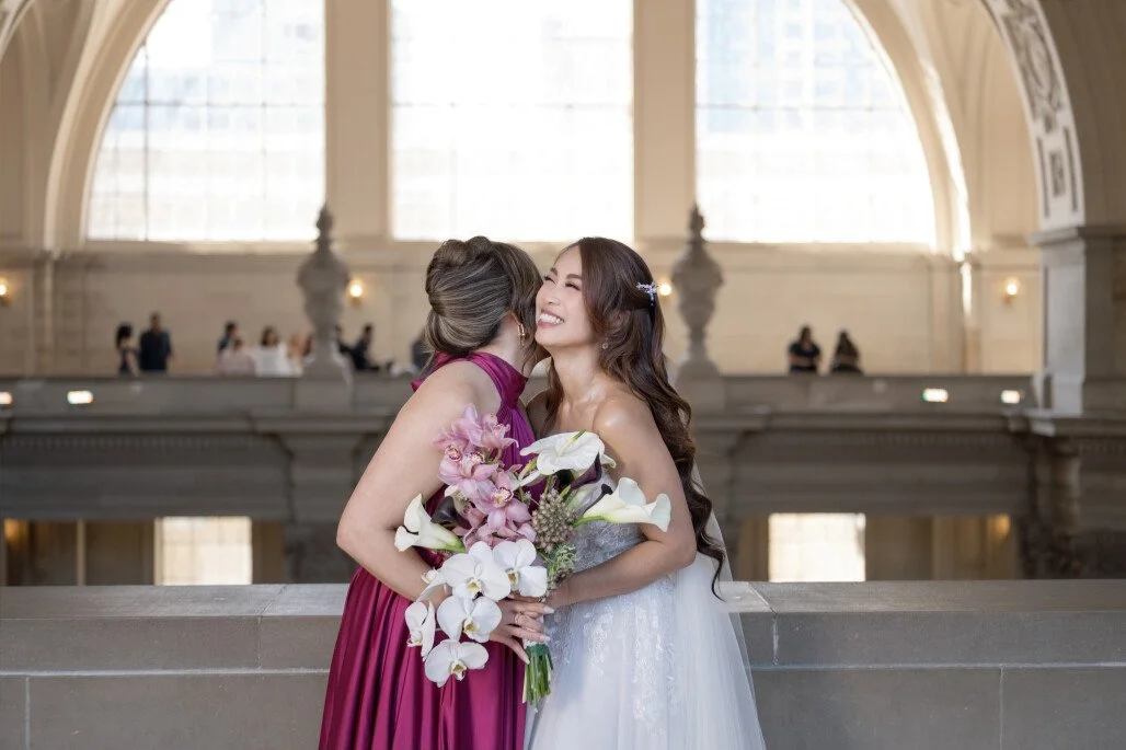Two women, one in a white wedding dress and the other in a pink gown, sharing a hug with the bride holding a bouquet of white and pink flowers inside a grand, well-lit building with large arched windows.