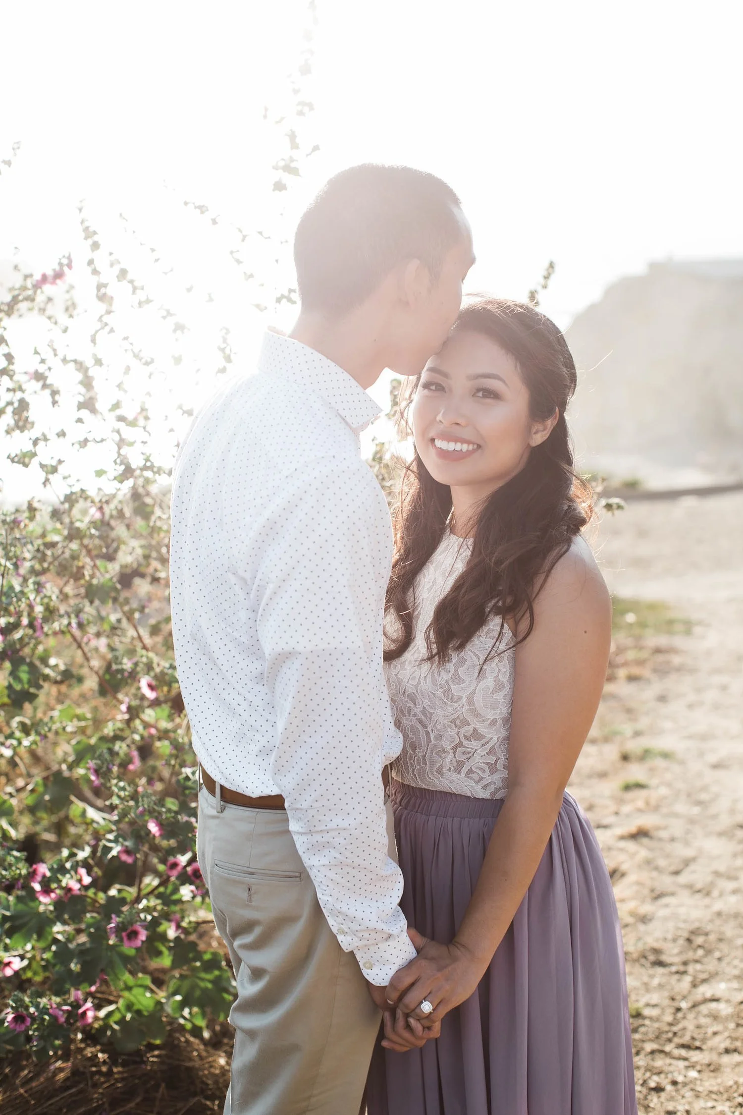 A young couple standing outdoors, holding hands, with the man gently kissing the woman's forehead. The woman is smiling warmly at the camera, wearing a lace top and a long skirt. The background features blooming flowers and bright sunlight.