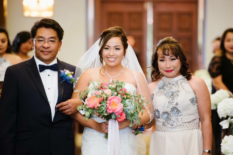 A bride in a white wedding dress holding a colorful bouquet, standing between a man in a tuxedo and a woman in a white dress with silver embellishments, indoors with wooden door behind them, smiling.