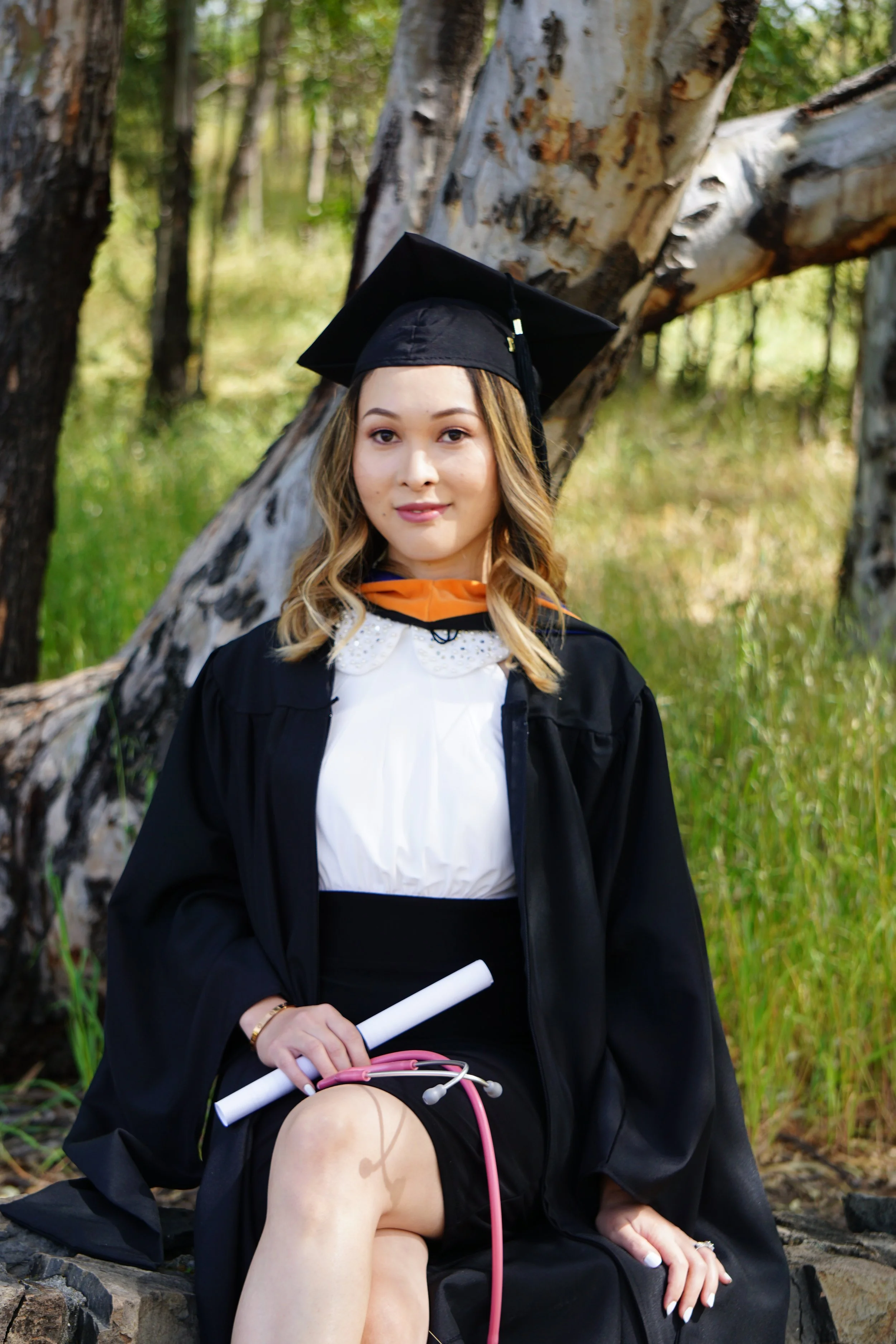 A woman in graduation attire sitting outdoors in front of a fallen tree, holding a rolled diploma and a pink stethoscope.
