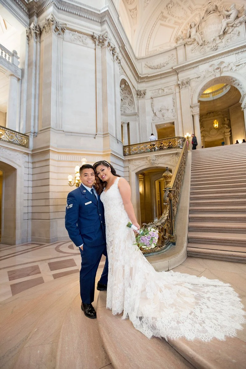 A bride in a white wedding dress holding a bouquet and a groom in a blue military uniform standing together on a grand staircase inside a historic building with ornate architectural details.