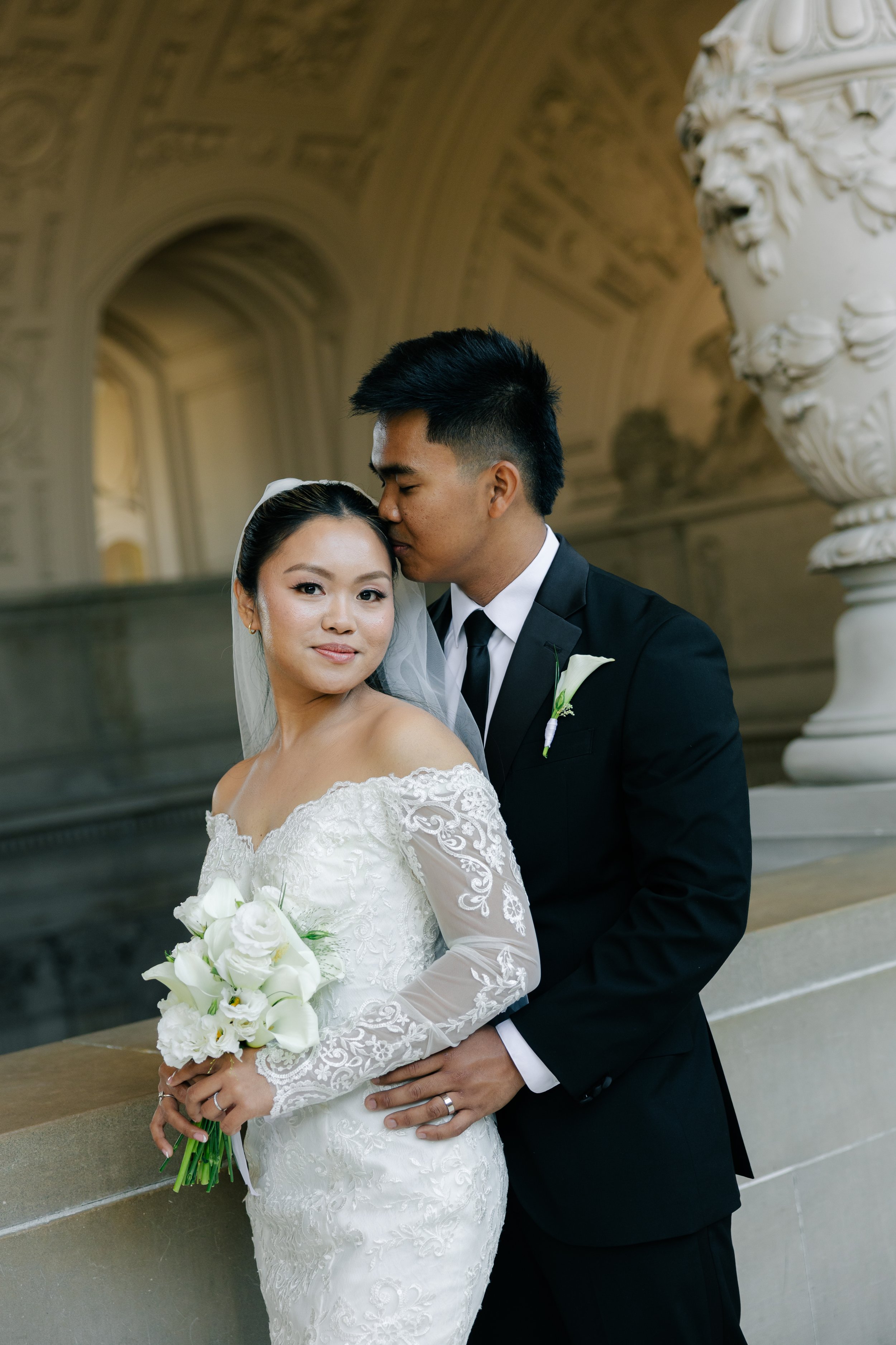 A newlywed couple stands in a historic architectural setting. The bride wears a white lace wedding dress and holds a bouquet of white flowers, while the groom wears a black suit and white shirt. The groom kisses the bride on the forehead as she smile