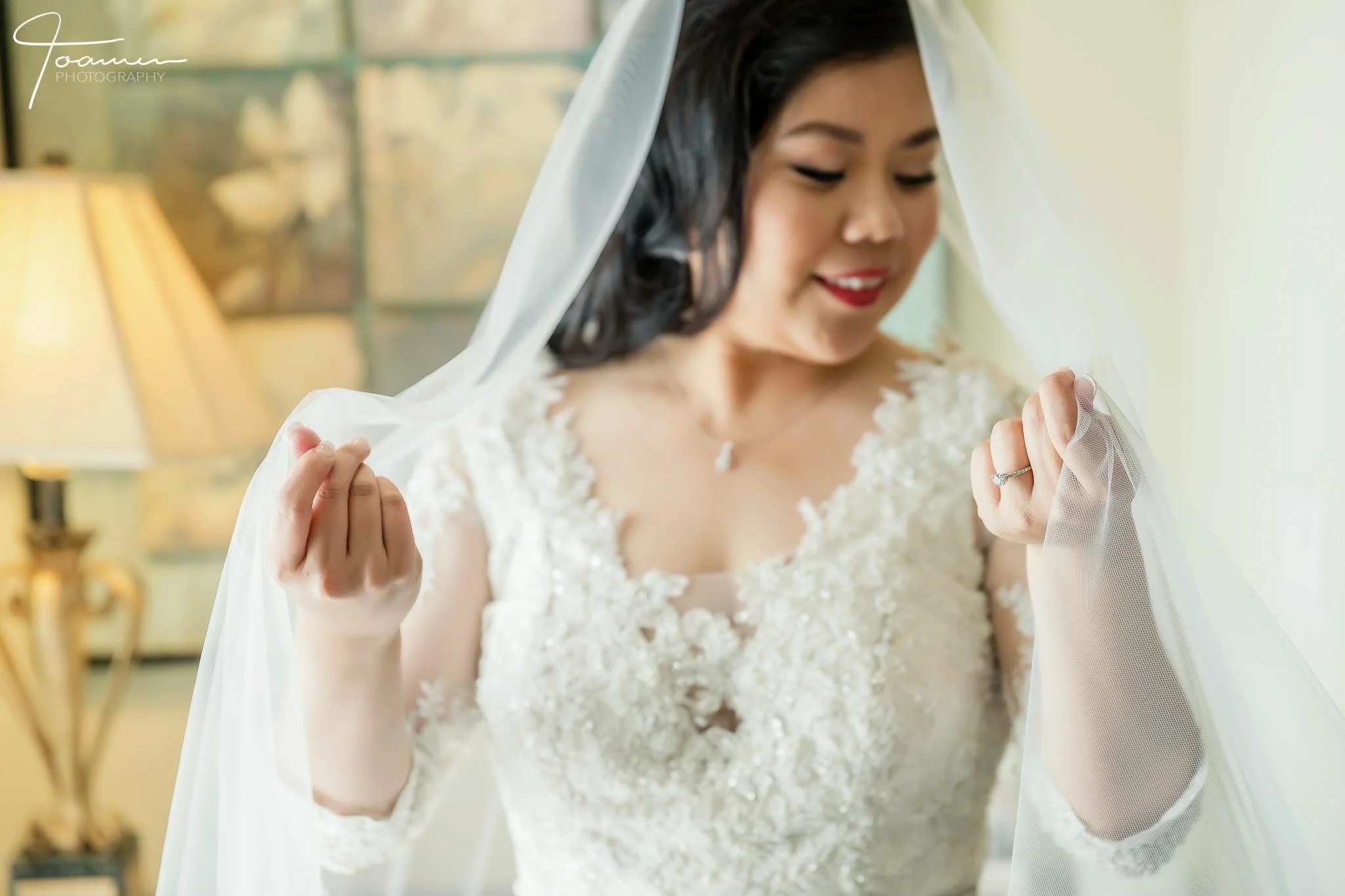 Bride with dark hair smiling and holding her wedding veil, wearing a lace wedding dress, in a warmly lit room.