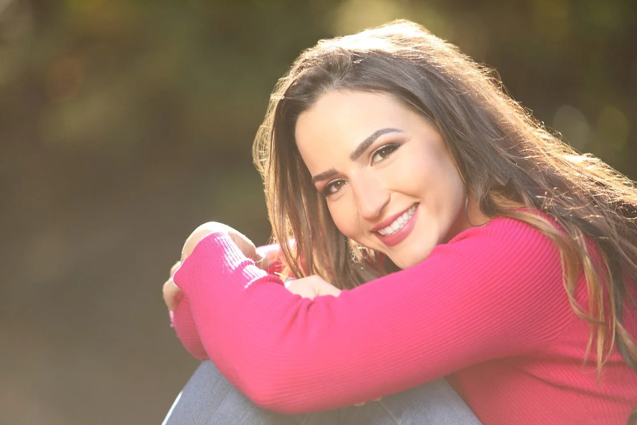 A young woman with long brown hair smiling and leaning on her knee outdoors during golden hour, wearing a pink long-sleeve shirt.