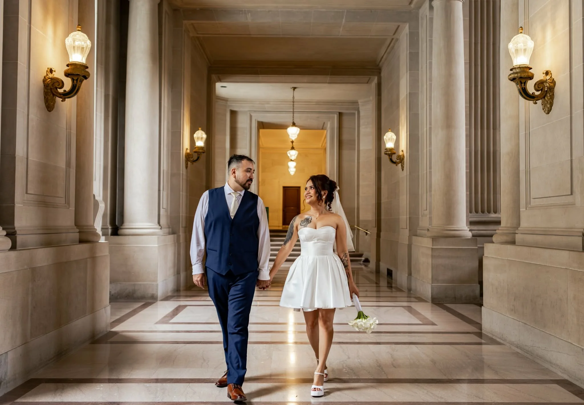 A couple dressed in wedding attire walking hand in hand in a grand, marble hall with tall columns and ornate wall sconces.