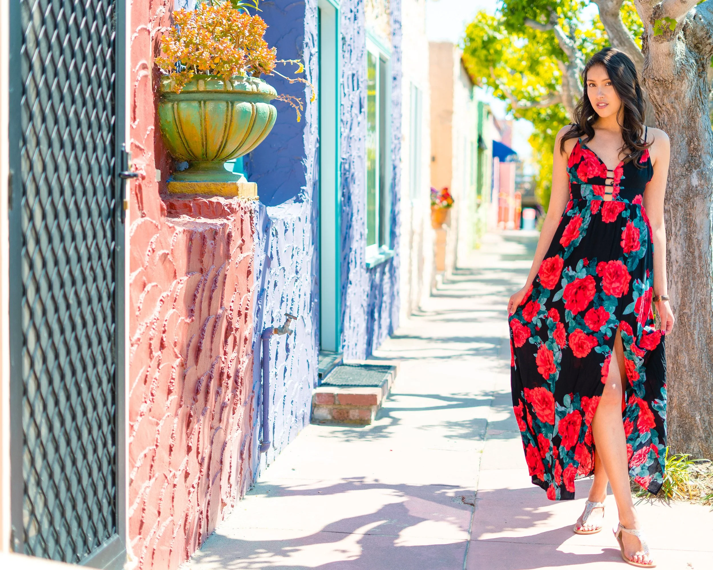 A woman walking on a colorful street with pastel pink, blue, and yellow houses, wearing a black dress with red roses and a thigh-high slit, with trees lining the sidewalk.