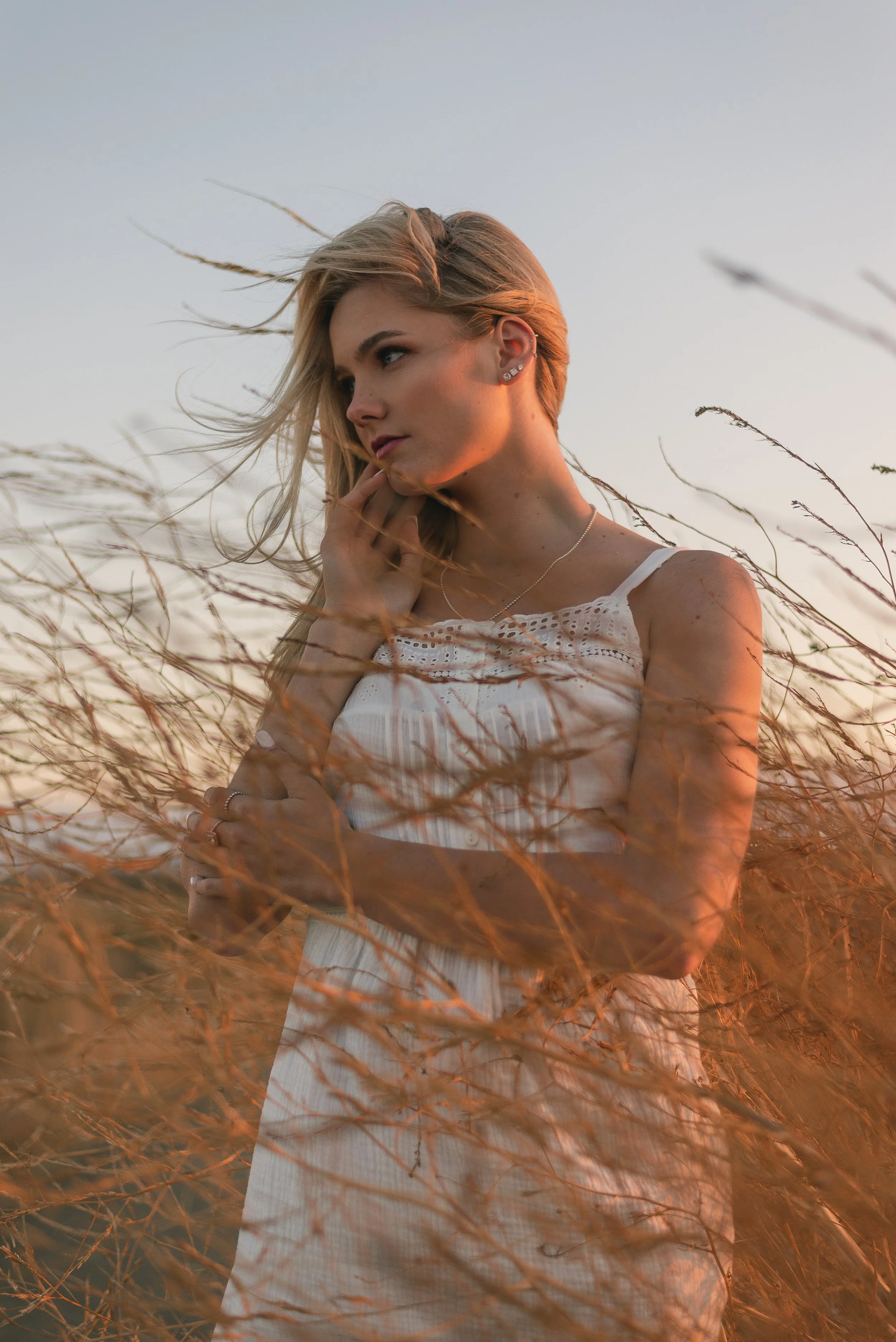 A young woman with blonde hair stands in a field of tall, dry grass at sunset, wearing a white sleeveless dress and looking thoughtful.