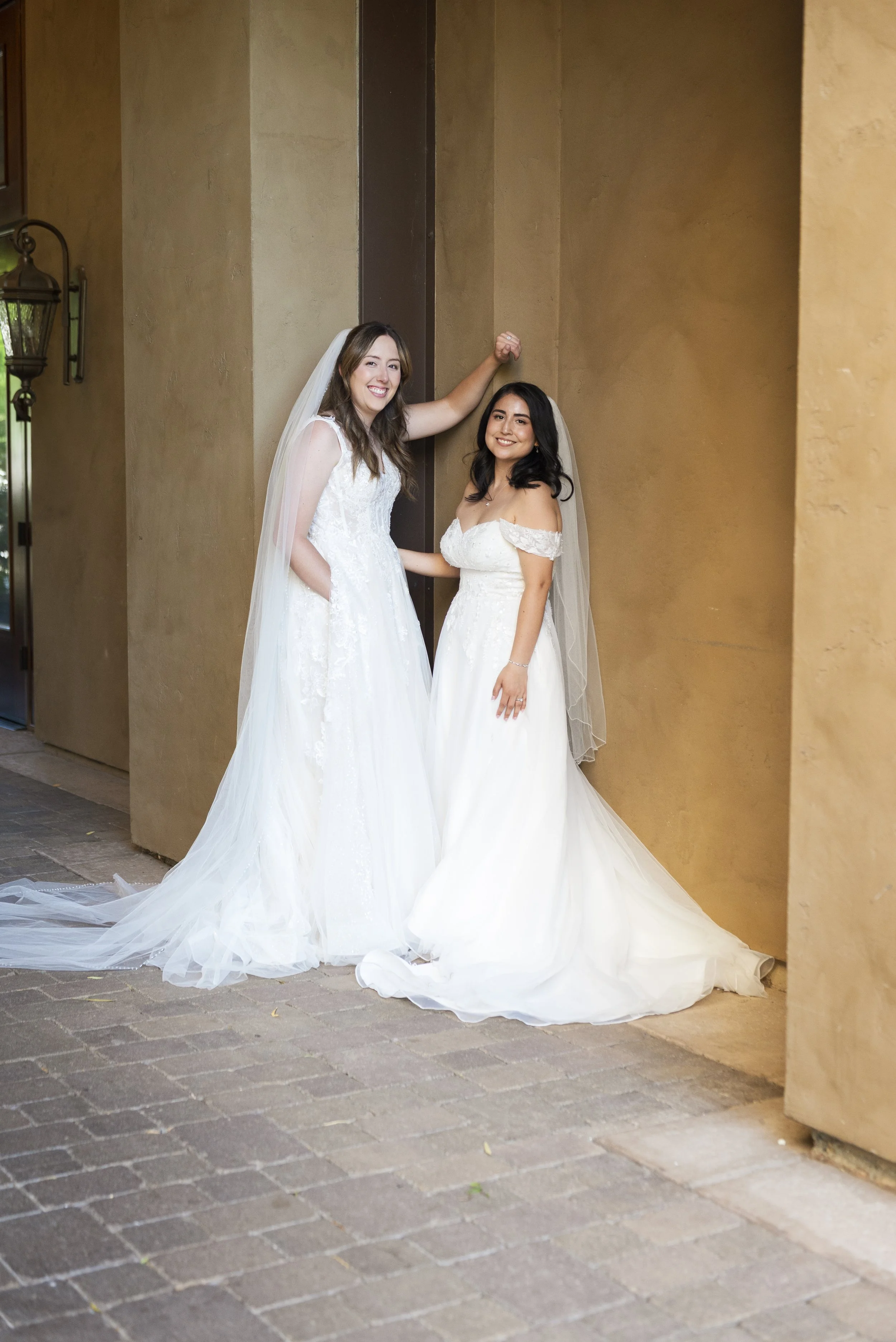 Two women in wedding dresses smiling and posing together in front of a beige wall.