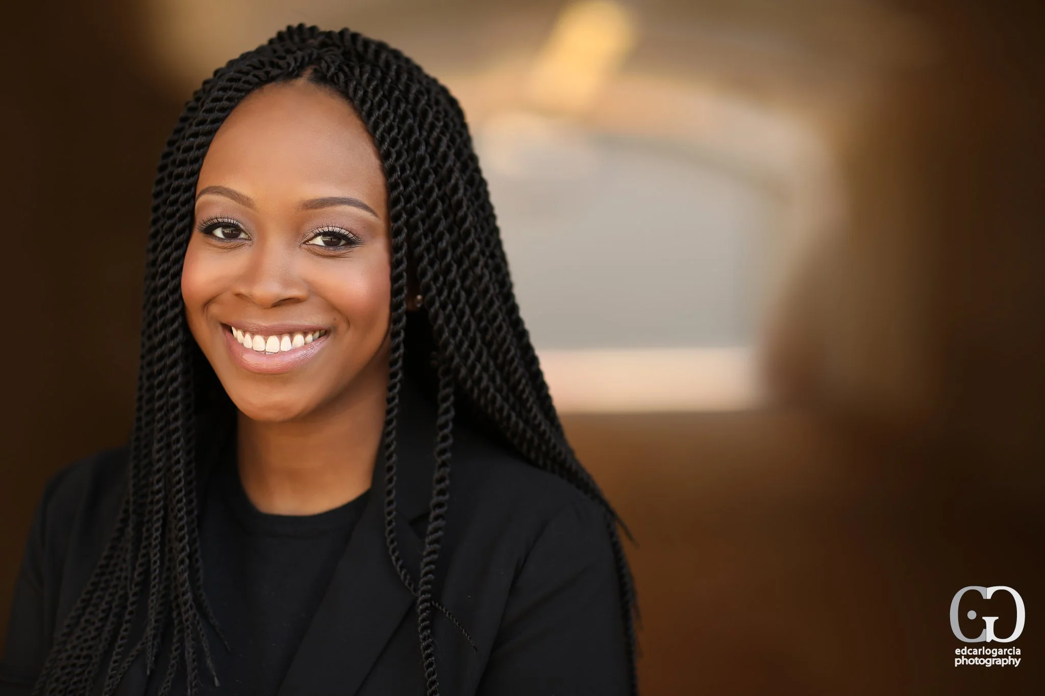 Portrait of a smiling African American woman with long braided hair wearing a black blazer against a blurred brown background.
