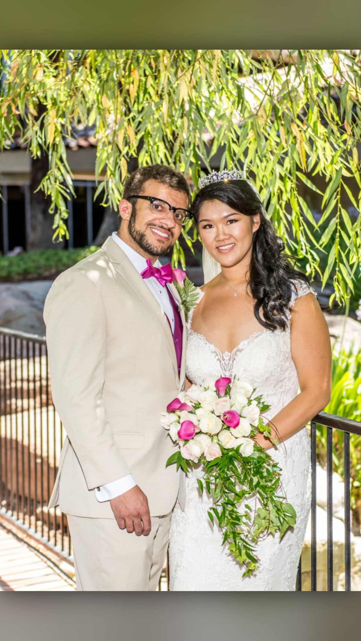 A bride and groom posing outdoors on their wedding day, with the bride holding a bouquet of white and pink flowers, and the groom wearing a beige suit with a pink bow tie, standing under a leafy tree.