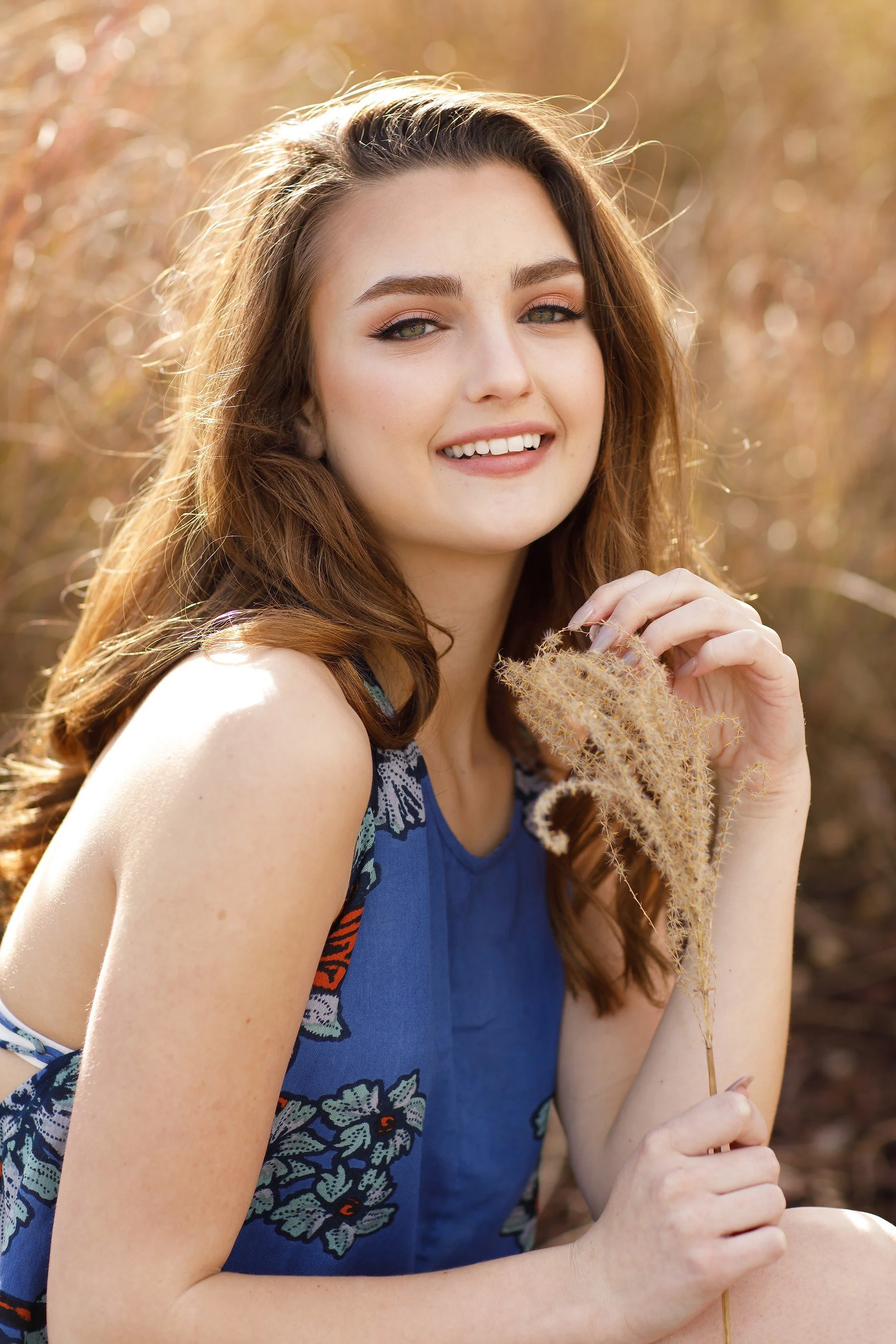 A young woman with long red hair holding a dried plant, outdoors on a sunny day with a blurred natural background.