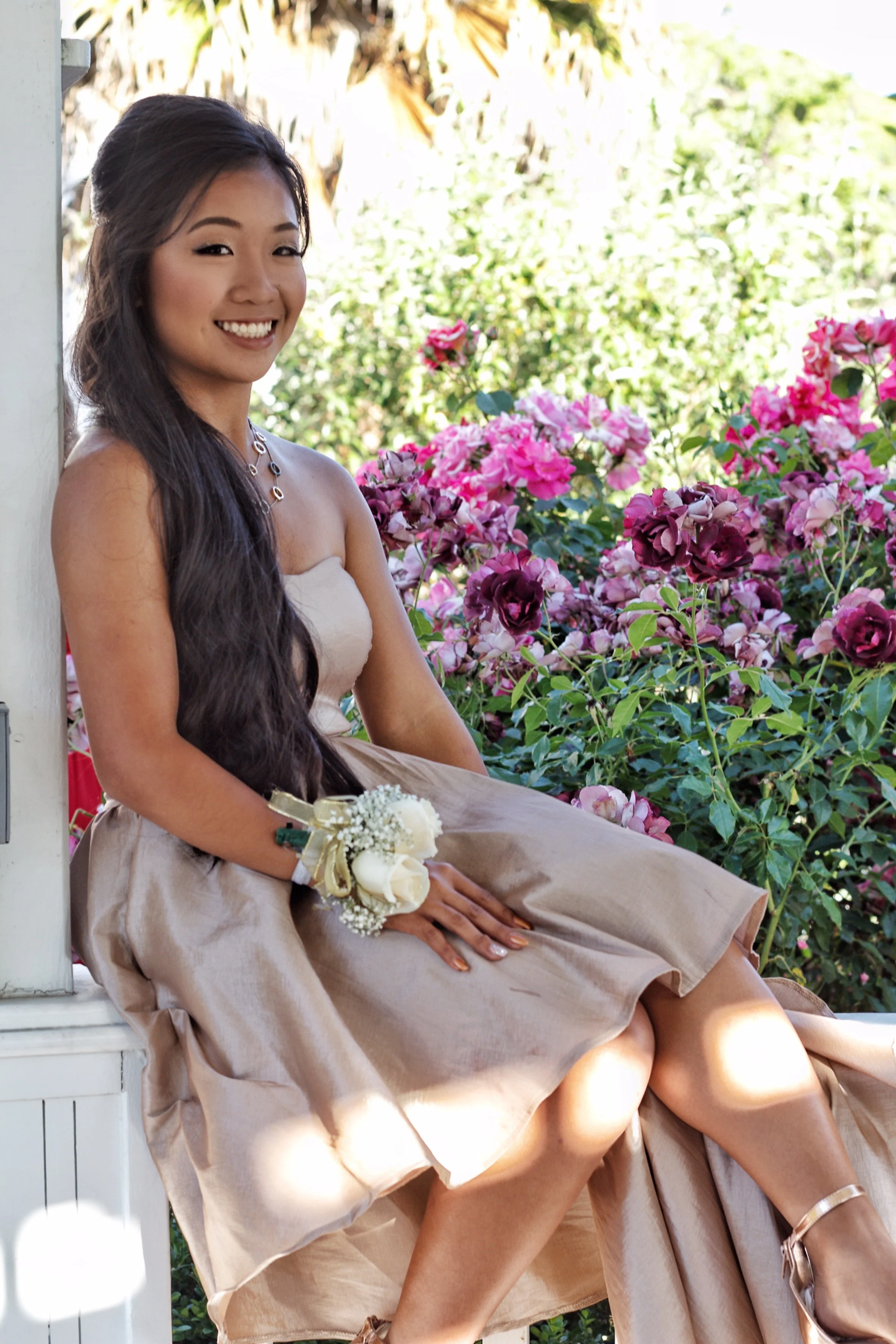 Young woman sitting outdoors near pink and purple flowers, wearing a strapless beige dress, with a corsage on her wrist, smiling at the camera.