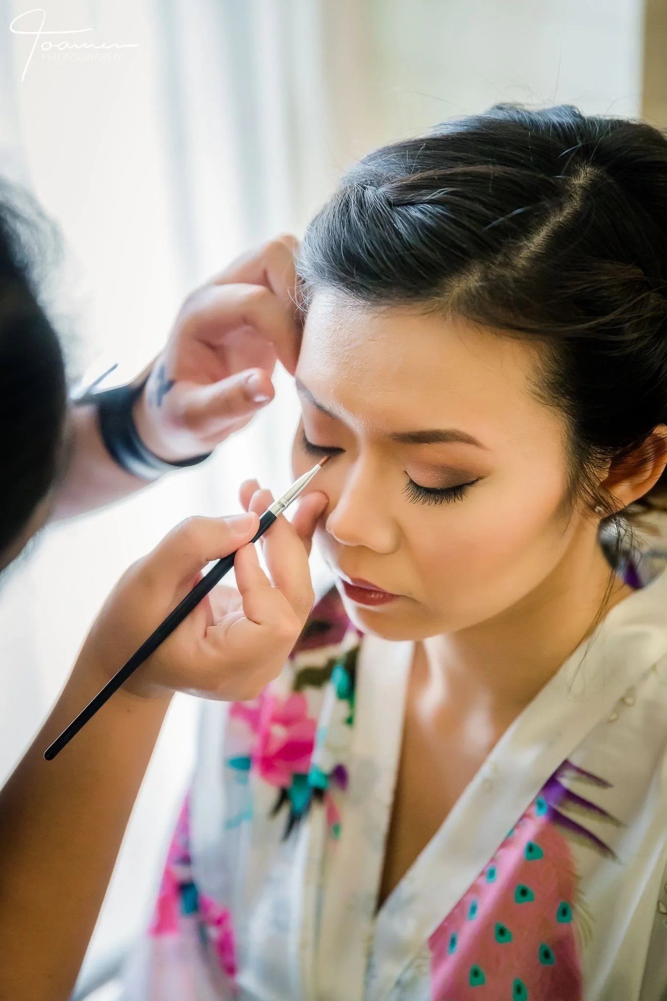 A woman with her eyes closed, wearing a floral robe, having her makeup done by a makeup artist who is applying eyeshadow or eyeliner near her eye with a brush.