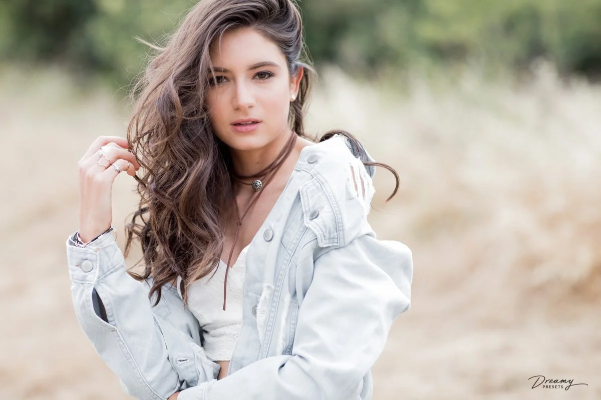 A young woman with long, wavy brown hair and light makeup posing outdoors with a blurred natural background, wearing a white top and a light-colored denim jacket.