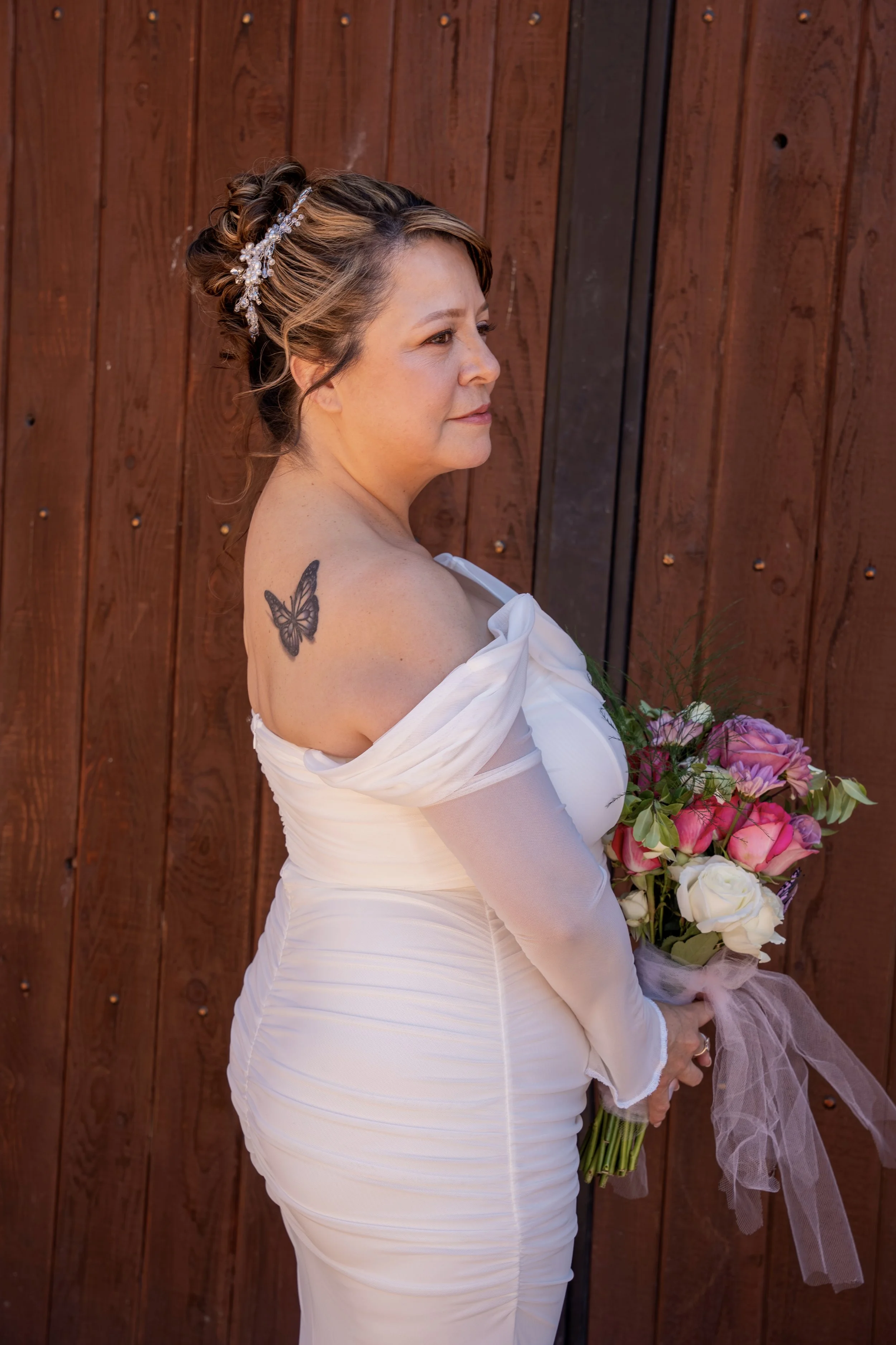 A woman in a white dress holding a bouquet of pink and white flowers, standing against a wooden wall.