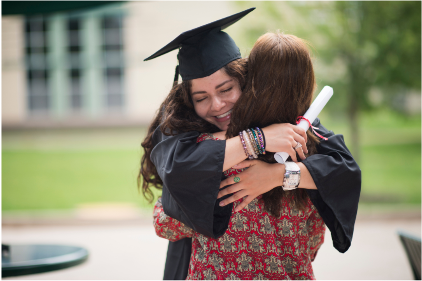 A young woman in a graduation cap and gown hugging another person outdoors on a grassy area.