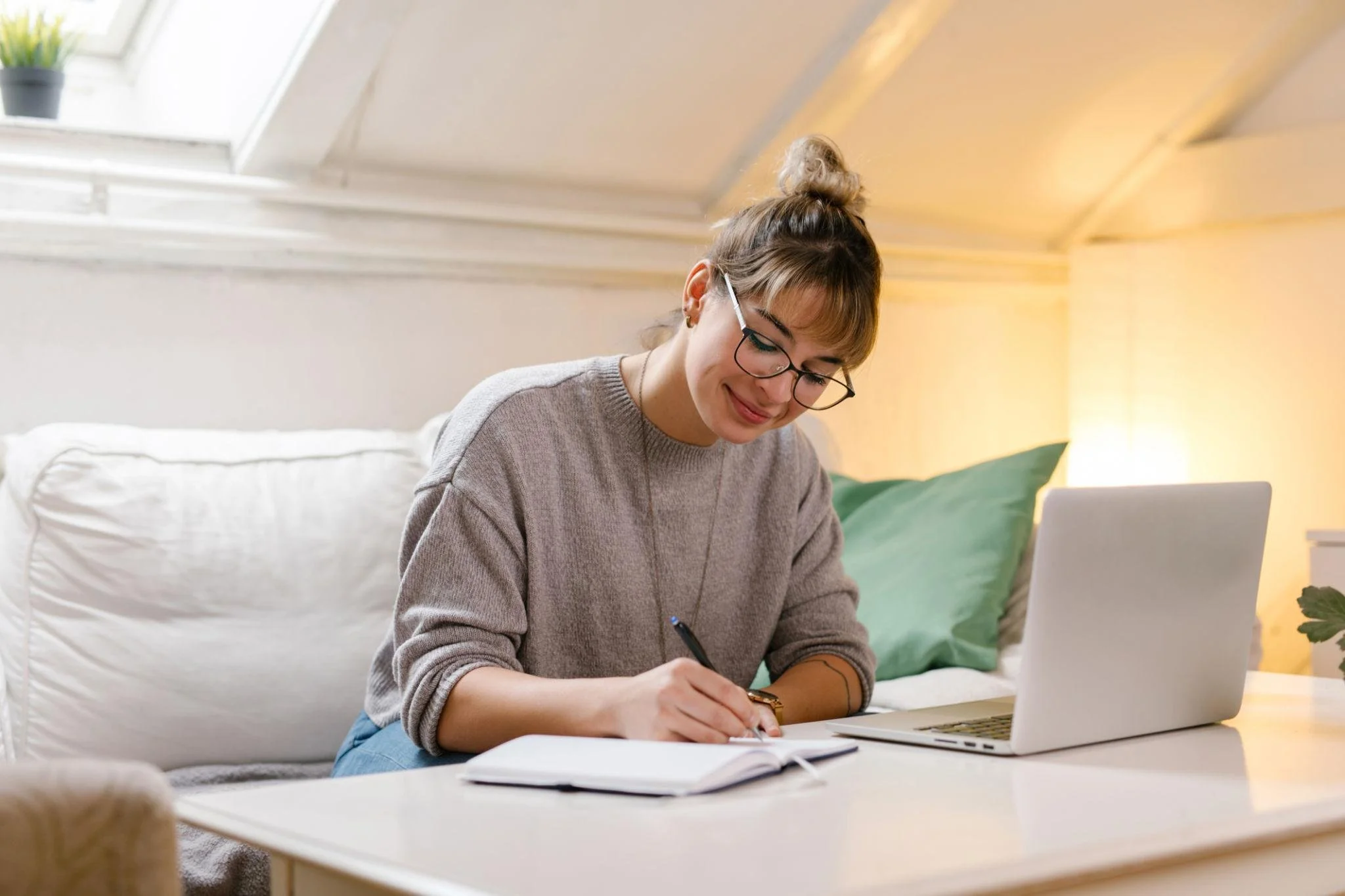 A young woman with glasses and a top knot hairstyle sitting on a beige sofa, writing in a notebook with a laptop in front of her in a cozy, well-lit room.