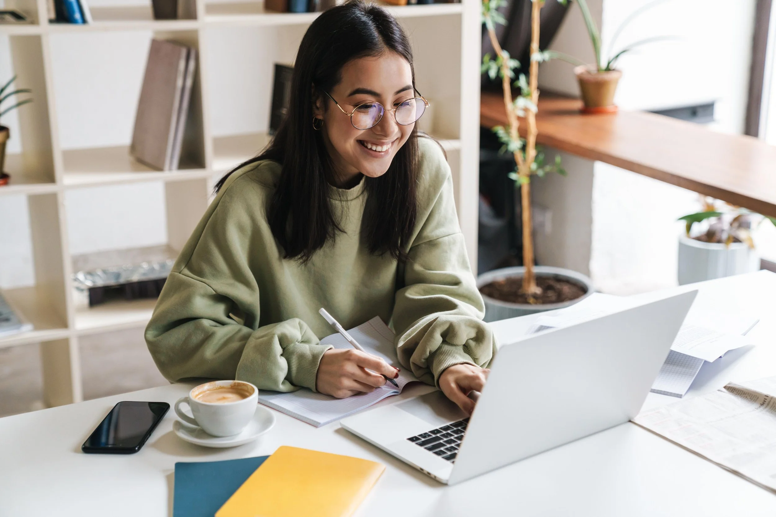 A young woman with long dark hair, glasses, and a green sweatshirt smiling while working on a laptop at a white desk in a bright, modern office space with plants and bookshelves.