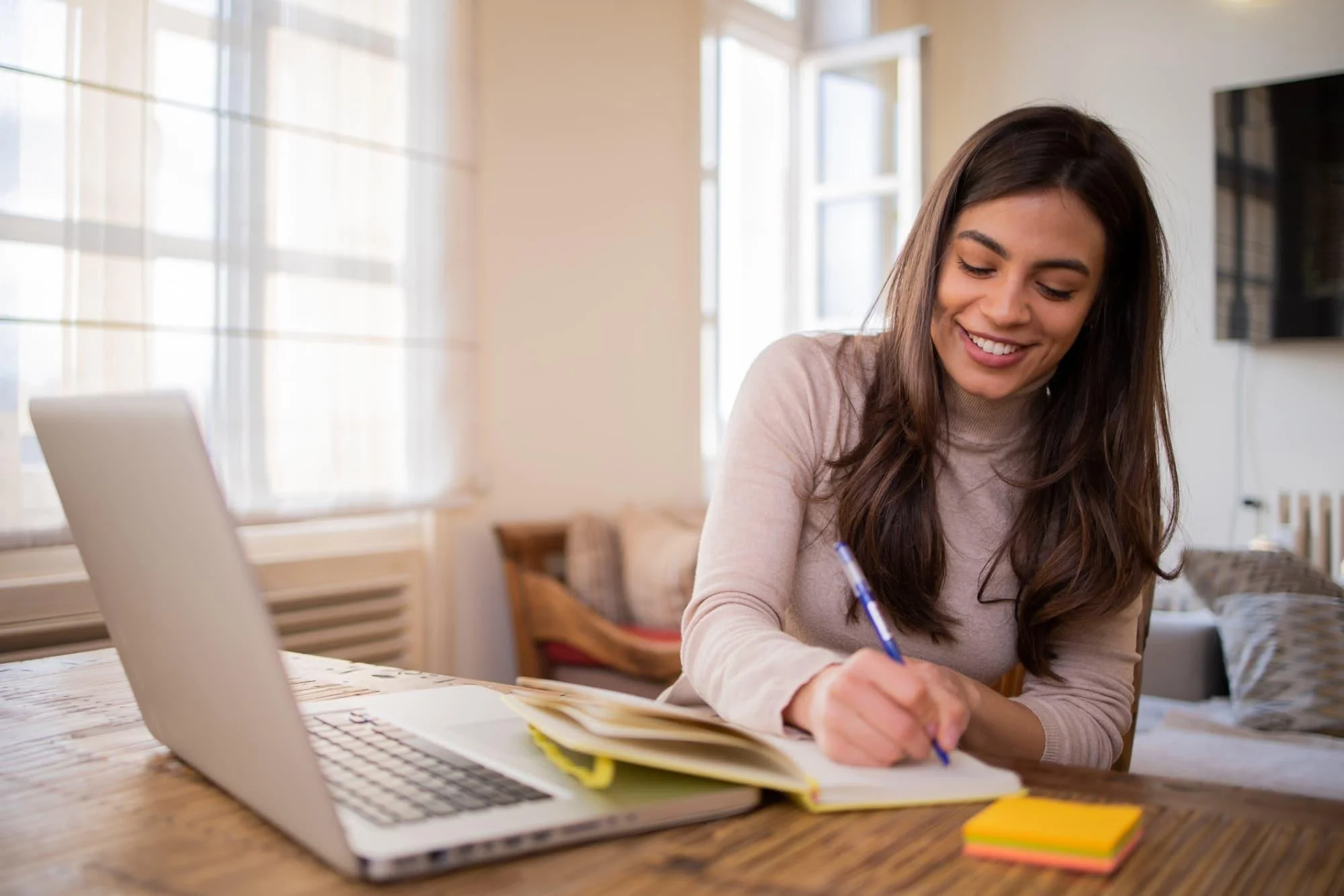 A young woman with long brown hair smiling while writing on paper at a wooden table in a bright room with large windows.
