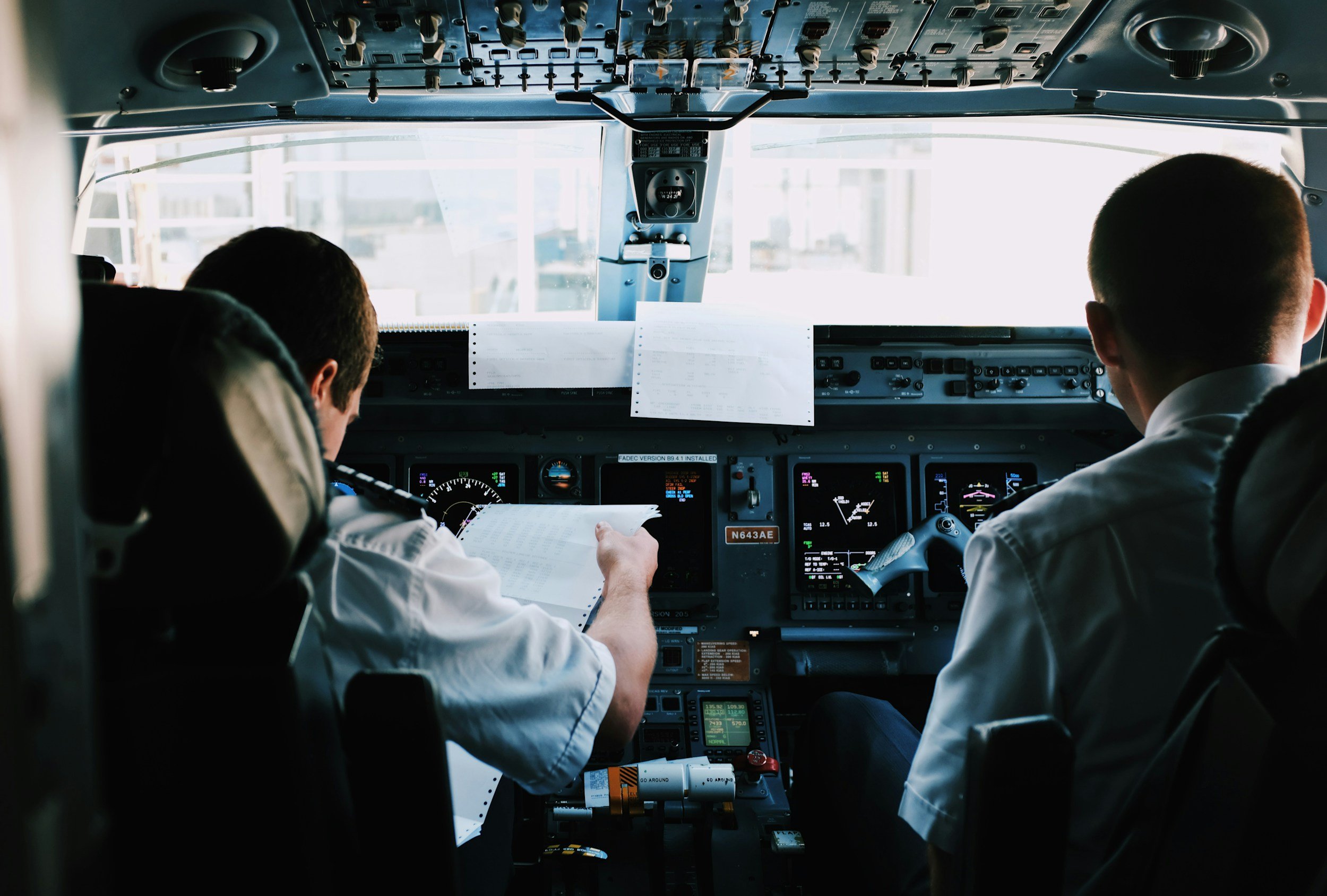 Two pilots in uniform seated in an airplane cockpit, reviewing documents and monitoring instruments.