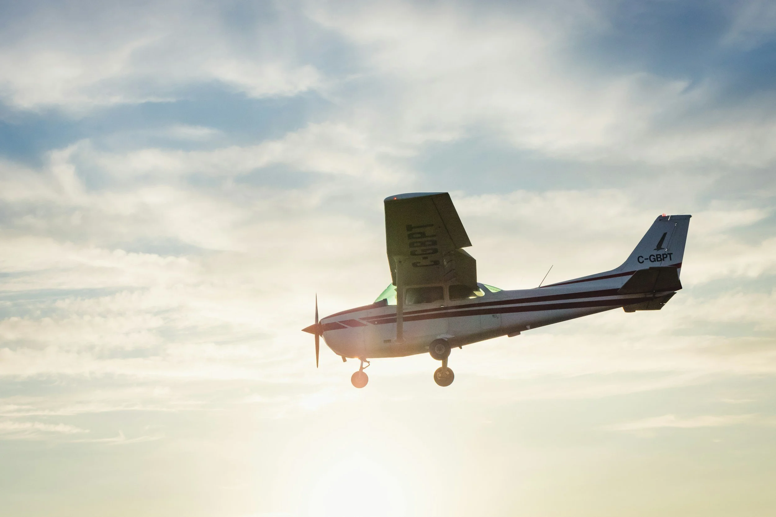 Small propeller airplane flying in a sky filled with scattered clouds, with the sun setting or rising in the background.