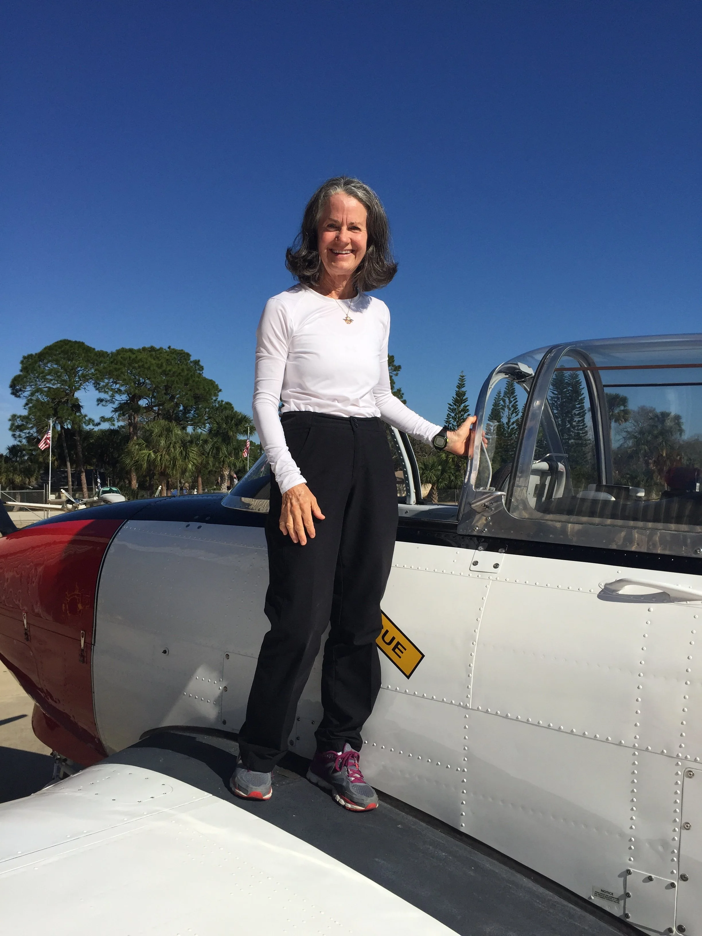 A woman with gray hair standing next to an aircraft, smiling and holding a door handle, wearing a white long-sleeve shirt, black pants, and gray sneakers.