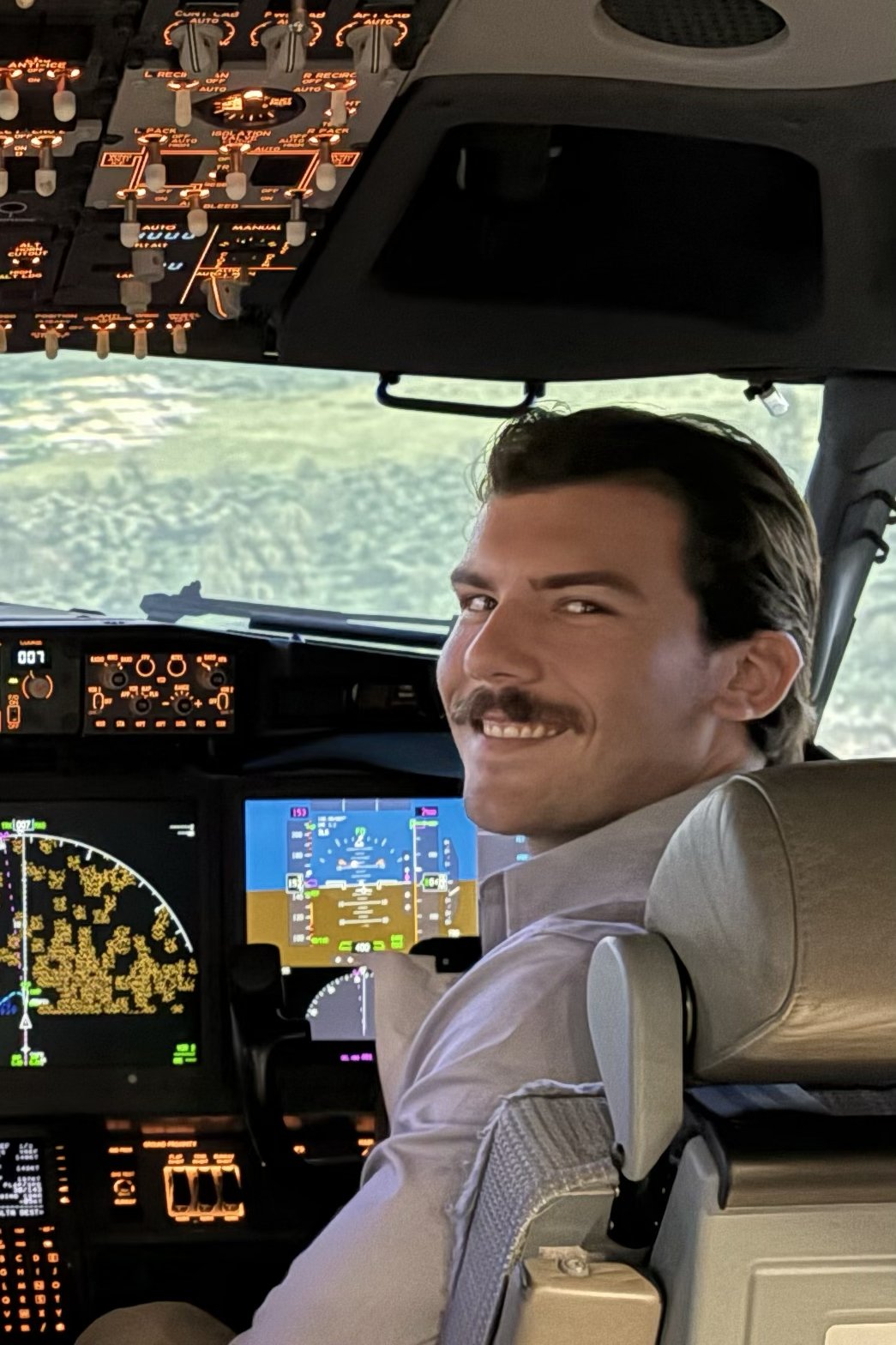 A smiling male pilot with dark hair and a mustache, sitting in the cockpit of an airplane with illuminated controls and screens, looking back over his shoulder.