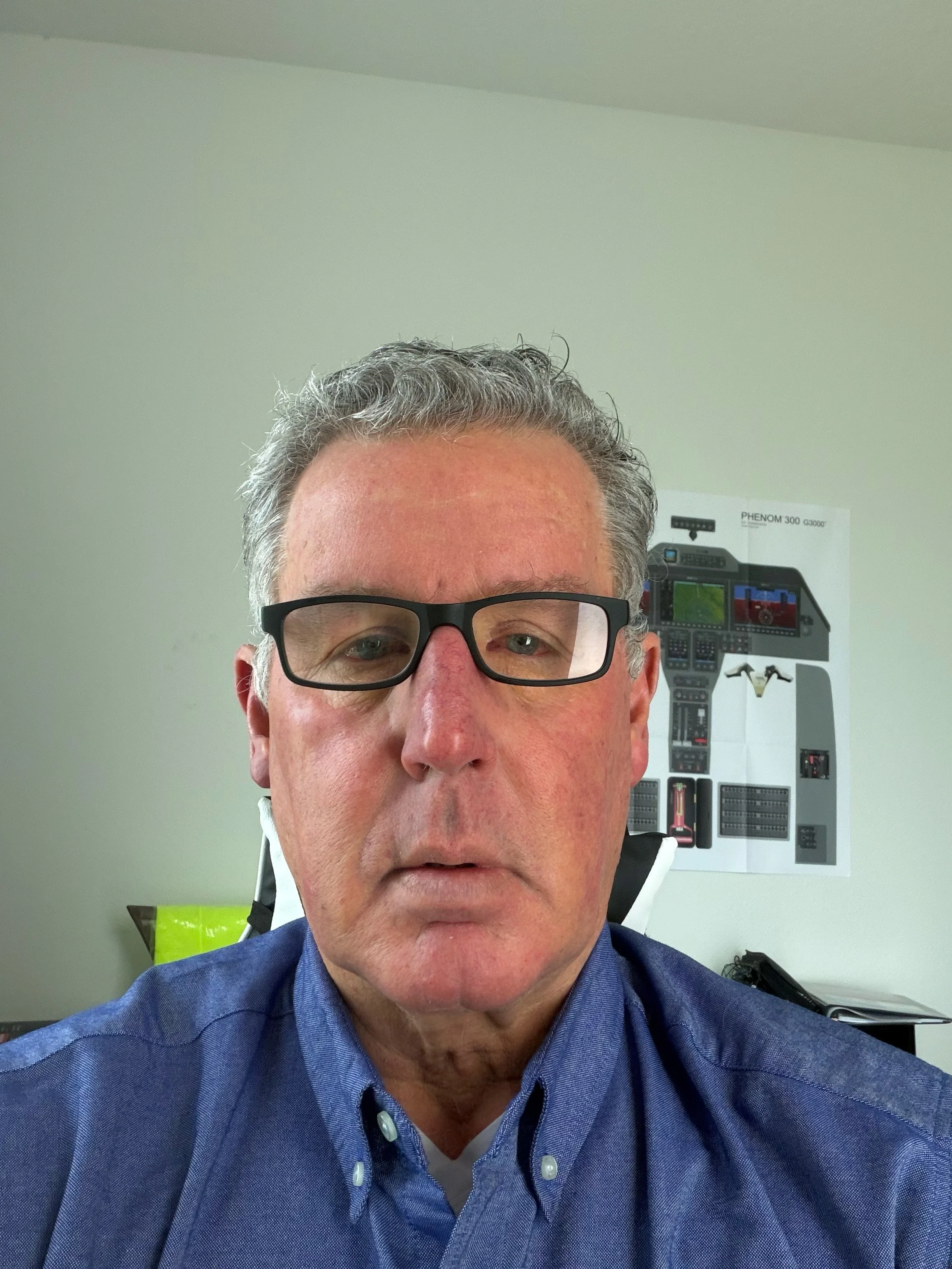 Close-up photo of a man with gray, curly hair and glasses, wearing a blue shirt, sitting in an office or medical setting with a poster and equipment in the background.