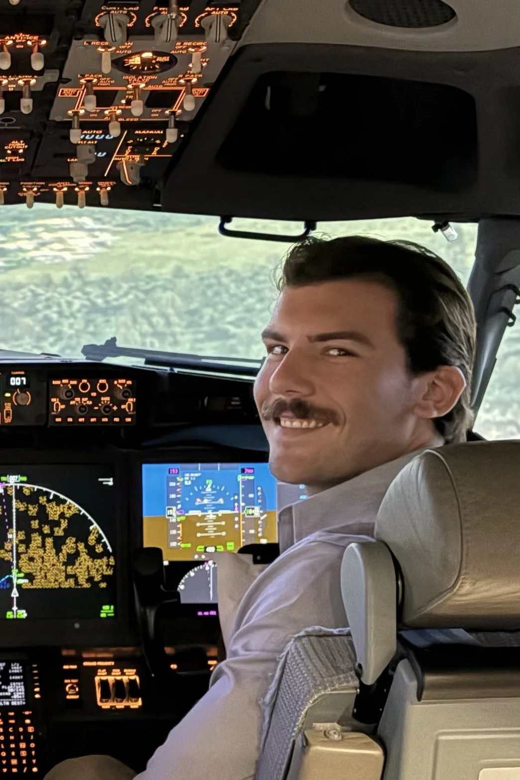 A smiling male pilot with dark hair and a mustache, seated in the cockpit of an airplane, with various control panels, screens, and instruments visible around him.