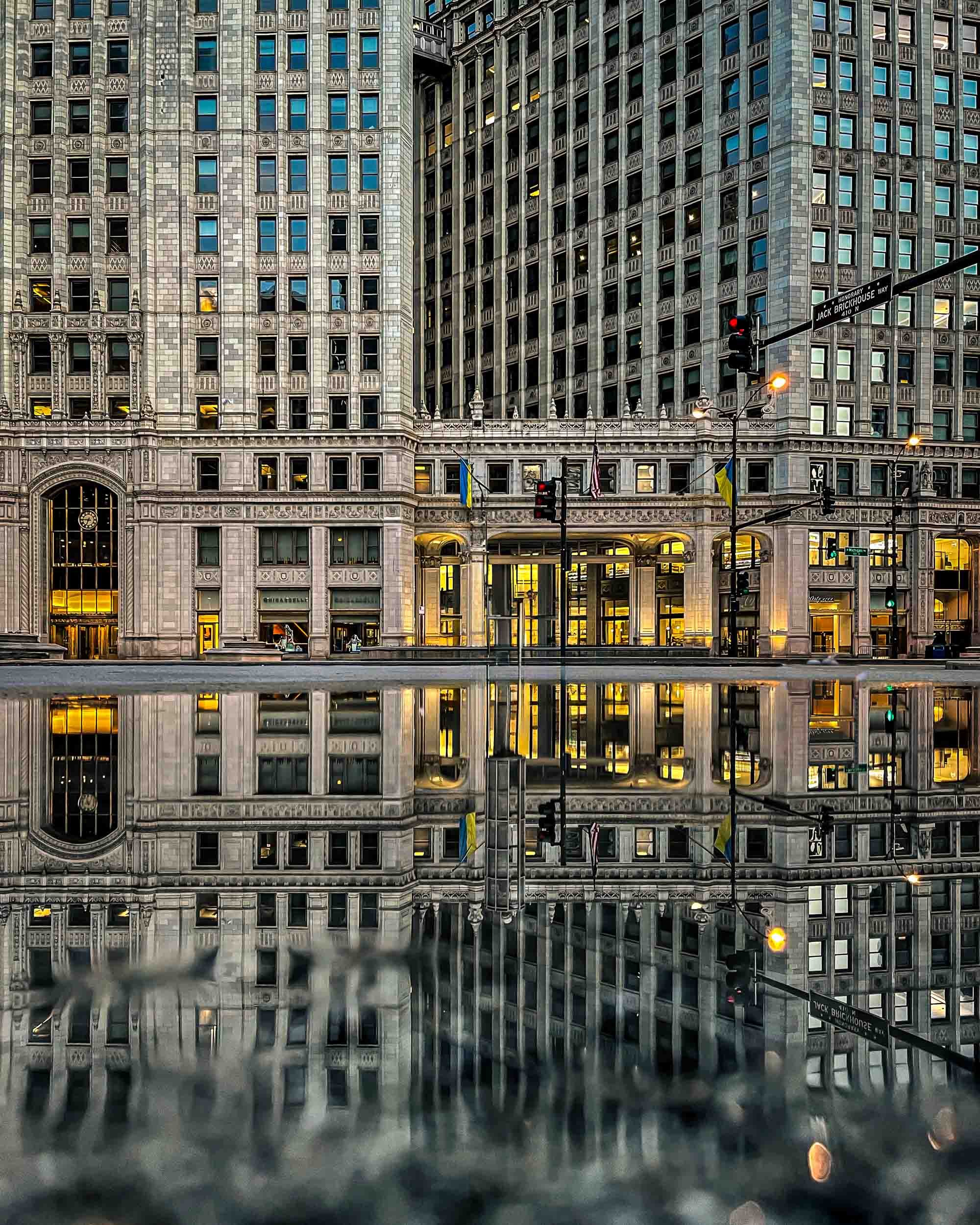 Reflection of Chicago office buildings with illuminated windows at dusk, seen on a wet street surface.