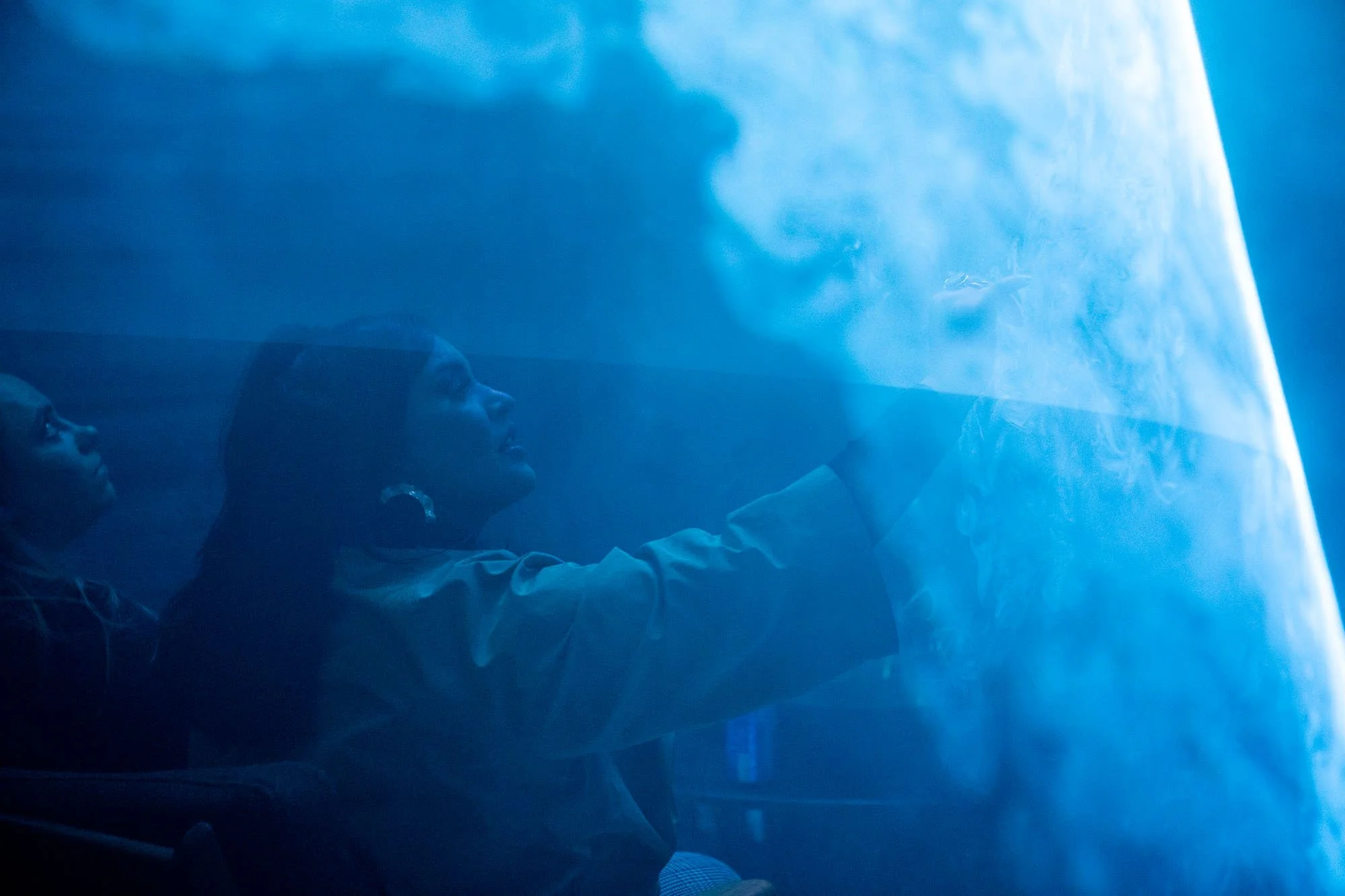 Two people, a man and a woman, face a wall made with a laser and haze. appearing to be a tunnel, with a blue hue and reflective surface they are interacting with.