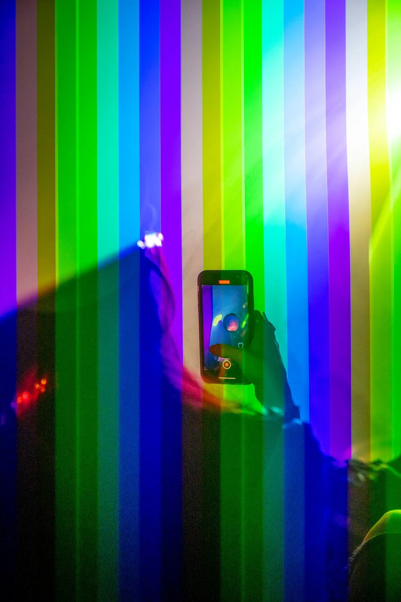 Colorful display with vertical LED strips in various colors, including purple, green, blue, and yellow. Someone is taking a photo of the display with a smartphone, capturing the reflection and the colorful lights.