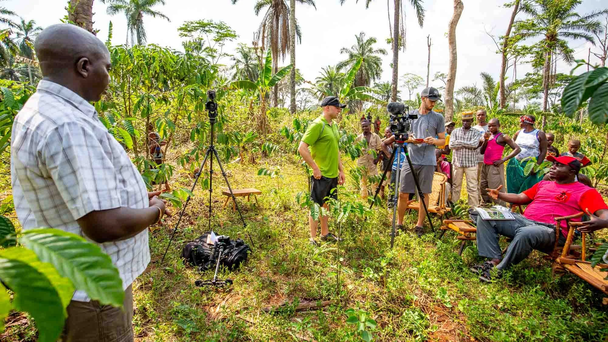 A group of people are gathered outdoors in a lush, green area with tall trees and dense foliage. Some individuals are setting up filming equipment, such as tripods and cameras, while others are sitting or standing, engaging in conversation or observation. The scene appears to be part of a documentary or interview being conducted in a natural setting.