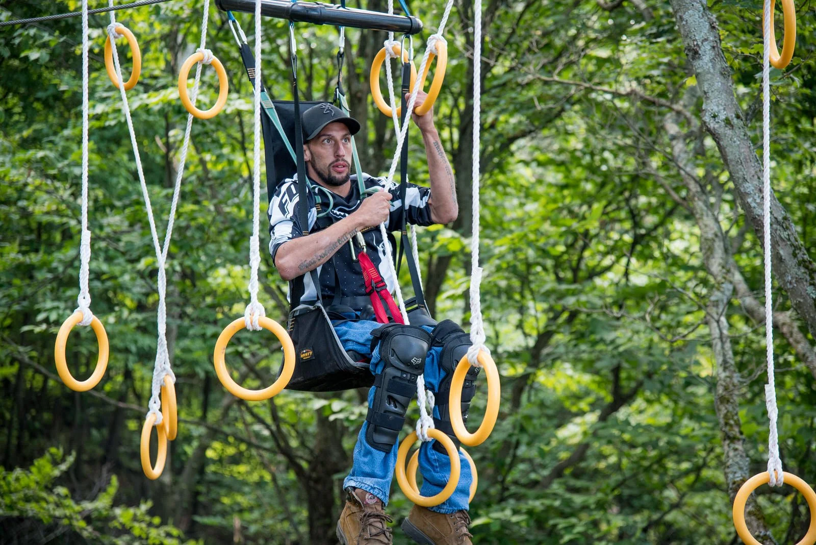 Man with disability in harness with climbing gear sitting on a swing suspended on rings in a forest, surrounded by green leaves on a high ropes course at The Root Farm.