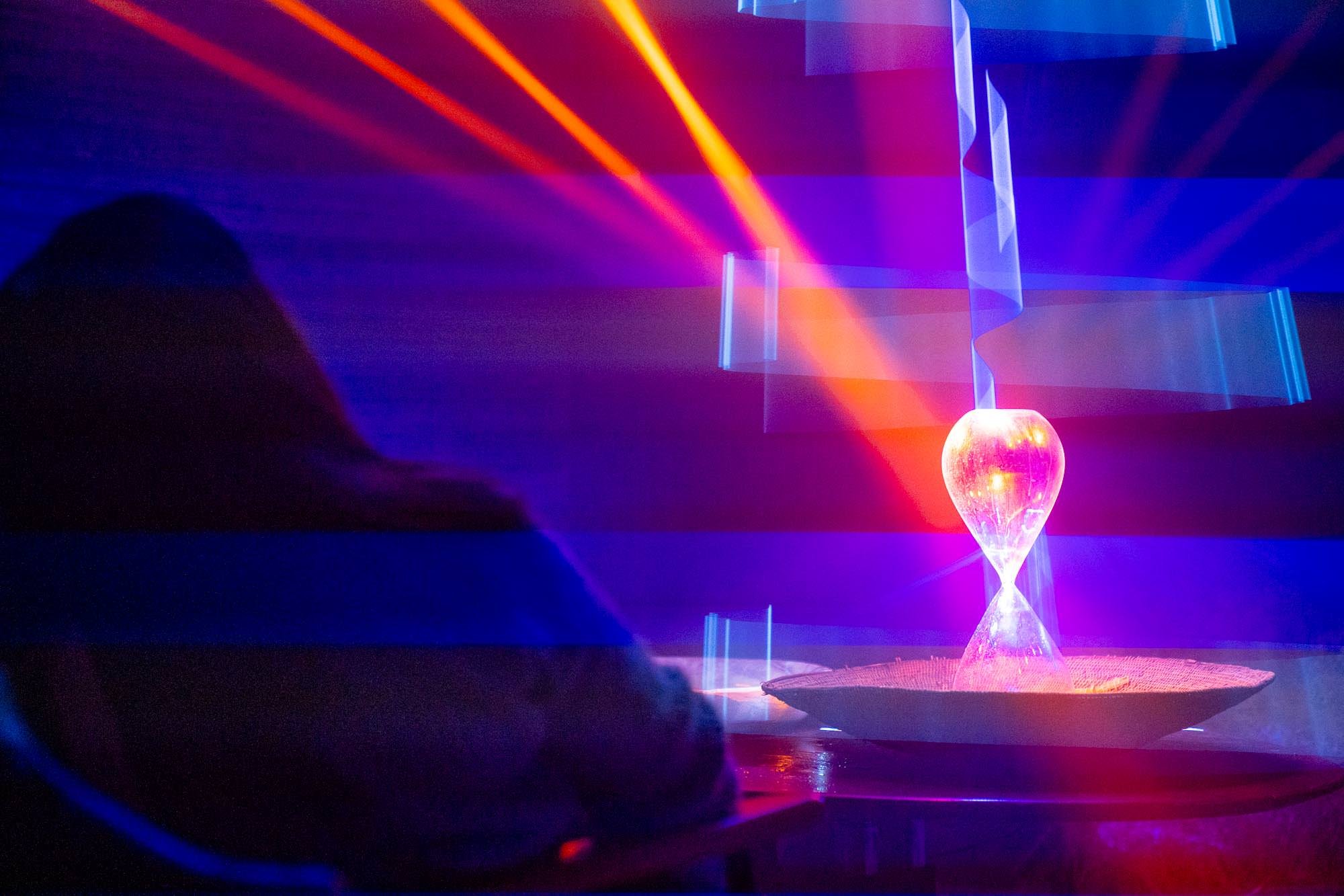 Colorful laser lights projected onto a dark background and an hourglass with glowing pink and purple colors.
