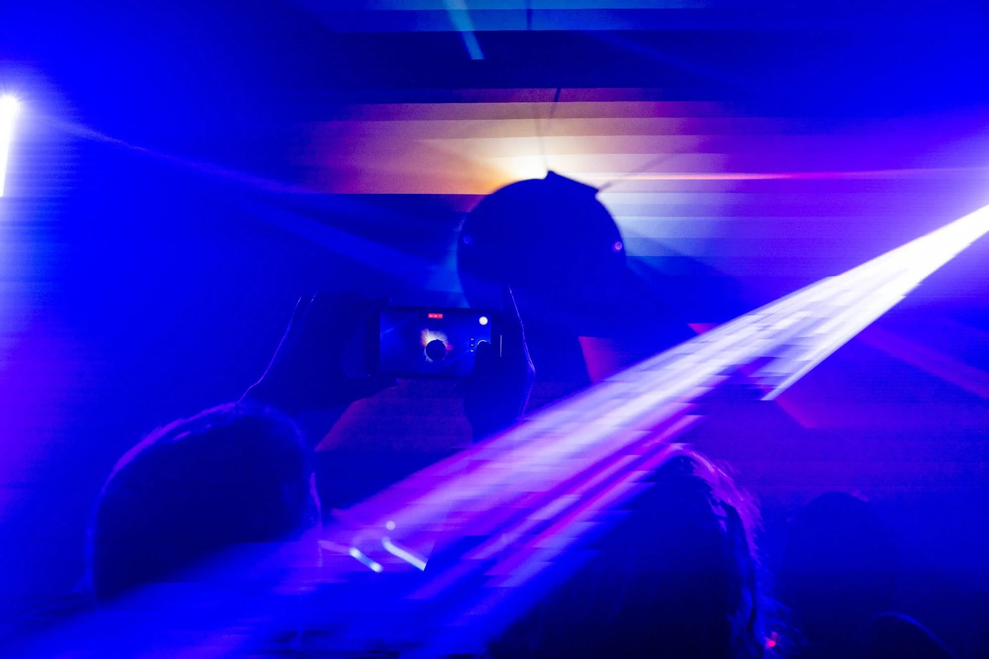 Person taking a photo of a laser light display with colorful blue, purple, and white lights in a dark room.