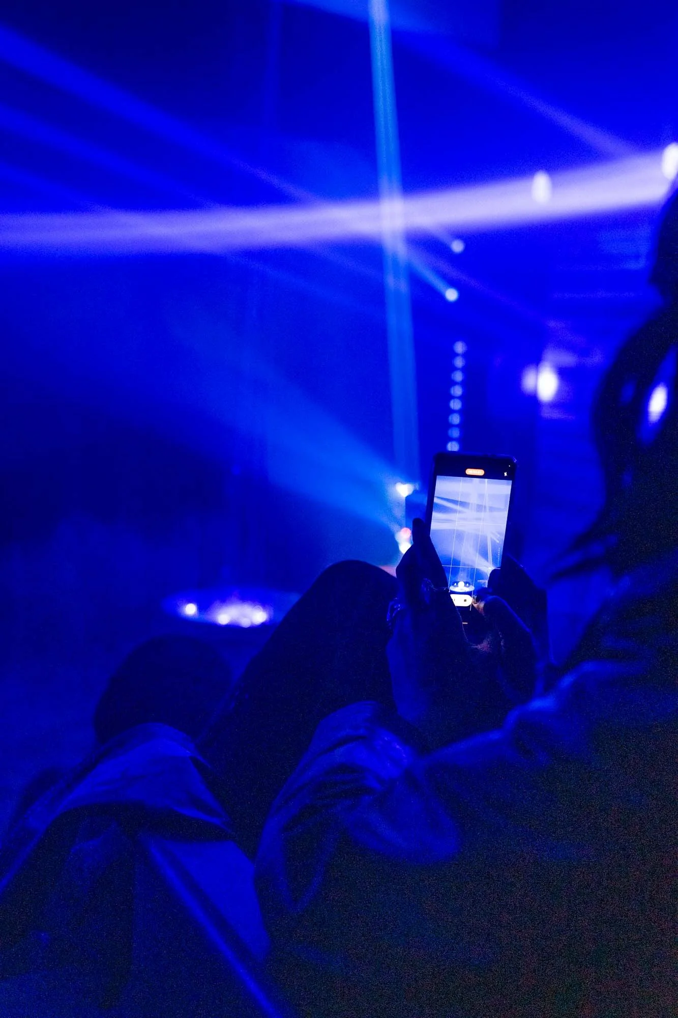 Person sitting in front of a stage with blue laser lights, using a smartphone to take a picture of the lights.