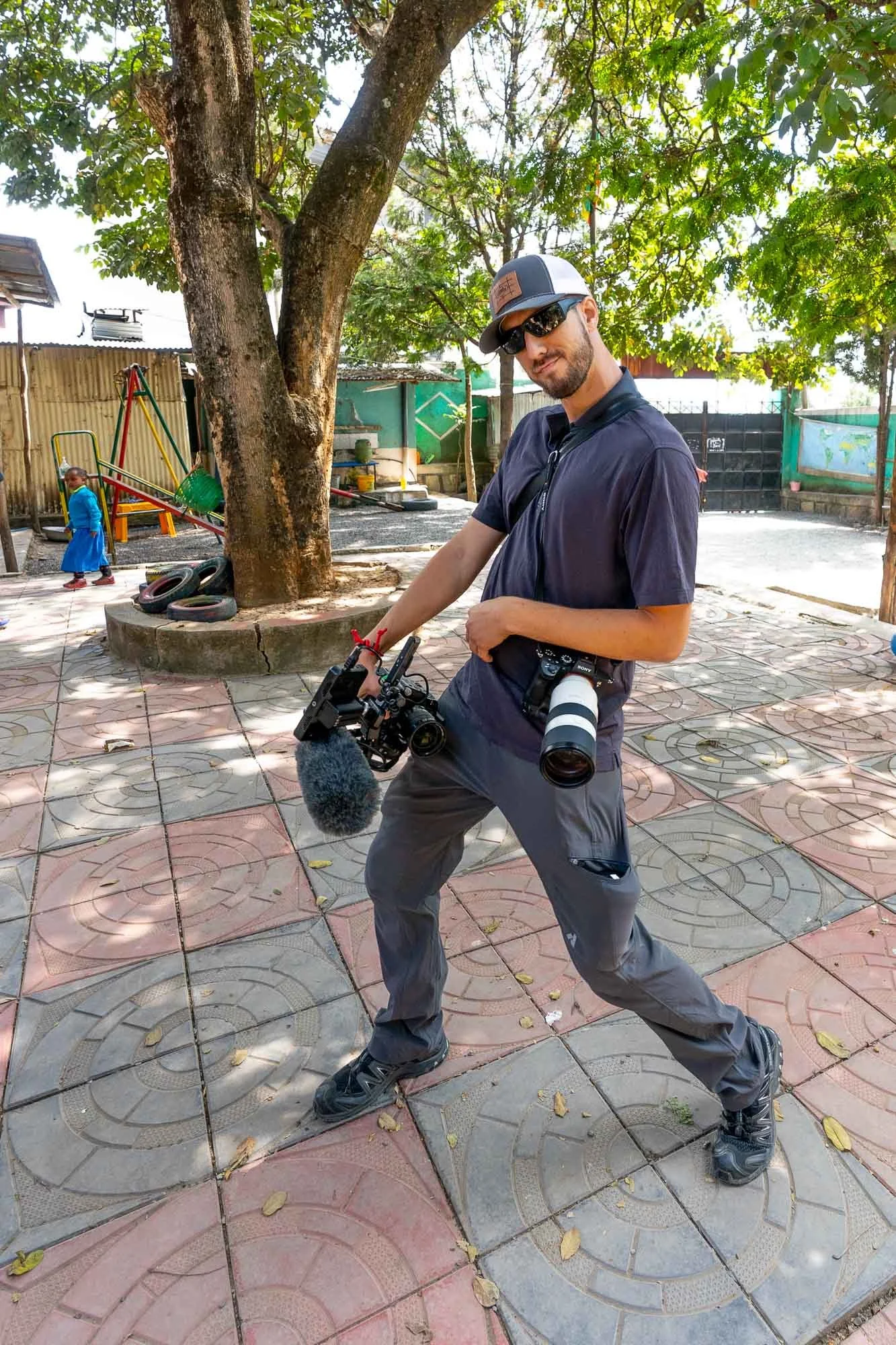 A man wearing sunglasses, a black T-shirt, gray pants, and a gray cap with a camera and a microphone attached, standing outdoors on a tiled surface with fallen leaves, a large tree, children playing on playground equipment, and a fence in the background.